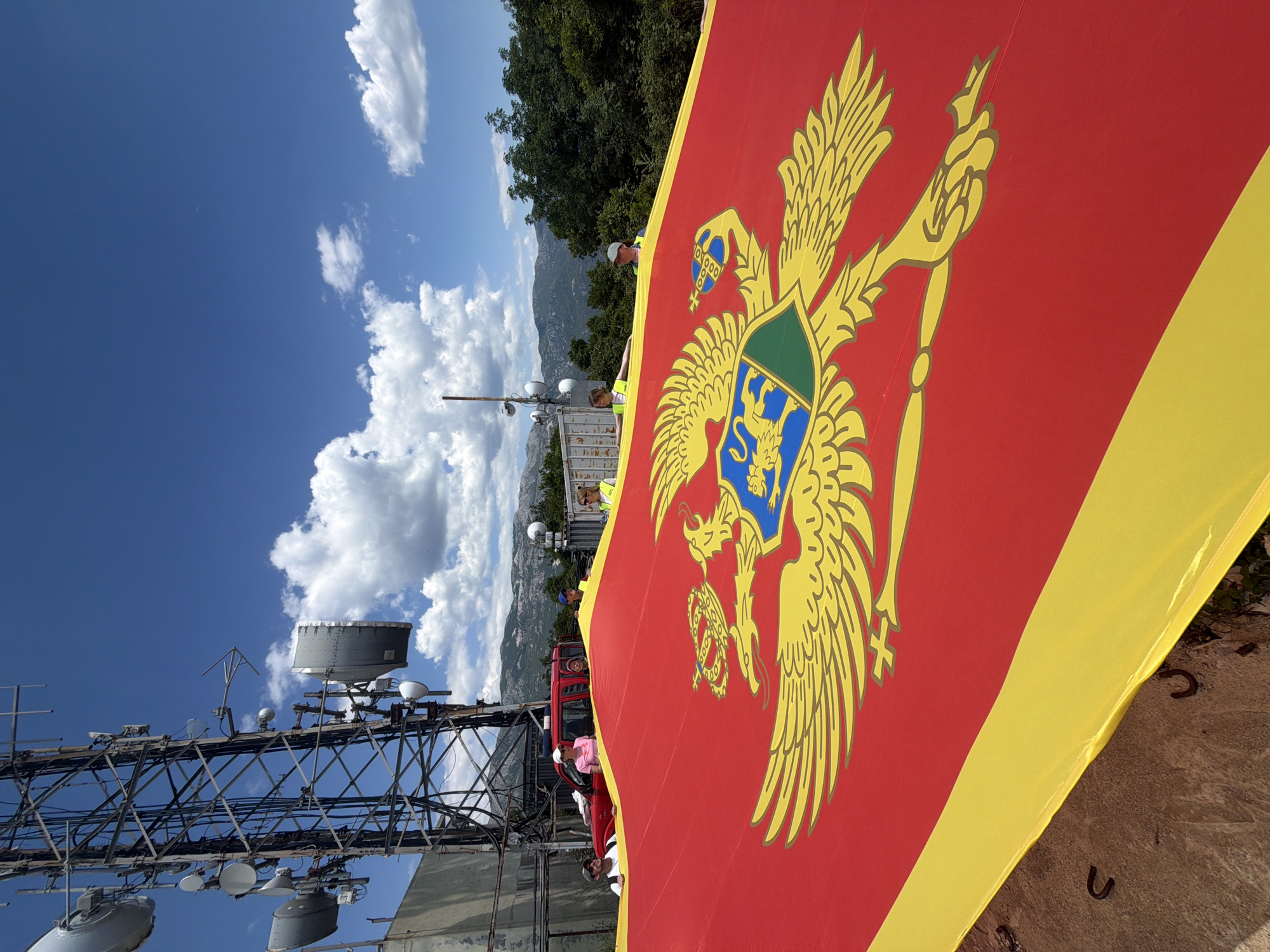 The image depicts the flag of Montenegro in the foreground, held by people whose presence is partially obscured by the flag itself. In the background, a radio tower stands on a landscape that includes visible mountains. The sky is clear blue with some scattered clouds. It is plausible that the image was taken from a high location overlooking Budva, Montenegro, given the mountainous landscape.