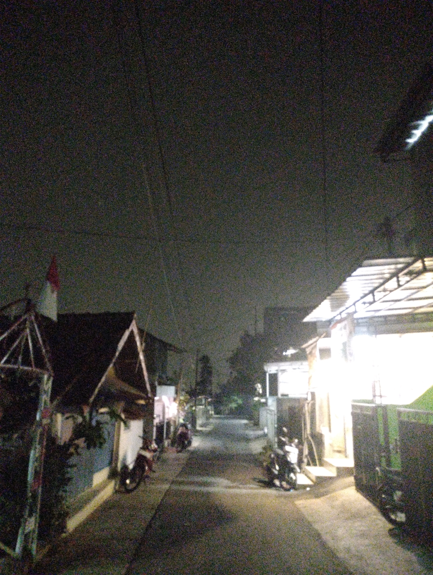 The image depicts a nighttime street scene in Cimenyan, Indonesia. The sky is dark and overcast, with visible noise in the image. On the left side of the frame, a house with a dark roof is visible, featuring a red and white flag on a pole. In front of the house, a metallic structure with diagonal supports is present. To the right, a brightly lit building with a corrugated metal roof extends across the frame. Several motorcycles are parked along the paved street. One motorcycle, a red and white scooter, is parked in front of the house on the left. Further down the street, a group of motorcycles is parked near the brightly lit building. The overall impression is that of a residential area at night, with minimal activity.