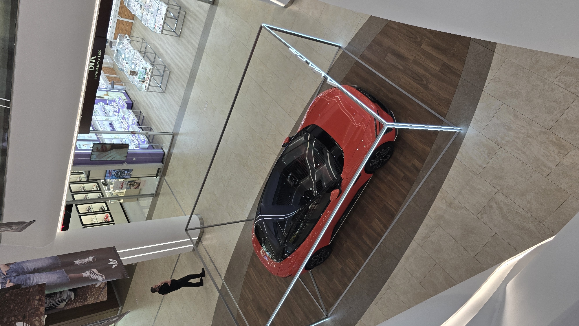 An aerial view captures a red sports car on display in the Atrium of the Ocean Plaza shopping mall in Kyiv, Ukraine. The car is positioned on a wooden platform and enclosed by a modern, illuminated glass and metal frame. To the left of the car, a man in a black shirt and pants stands talking on his phone, facing away from the camera. In the background, beyond the car display, is a jewelry store with display cases filled with watches and jewelry. The overall environment is that of a well-lit indoor shopping mall with tiled flooring.
