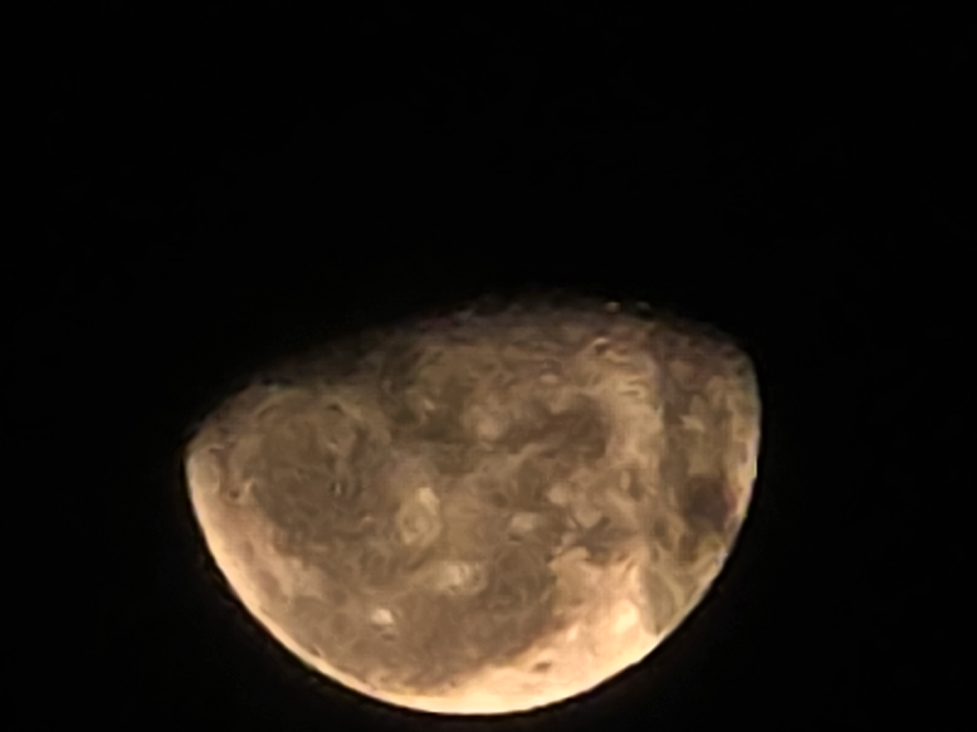 The image is a close-up of a celestial body, possibly a planet or moon, against a black background. The object is illuminated from the left, with a crescent shape visible. The surface of the celestial body appears textured with variations in color, ranging from light tan to a deeper brown. There are no discernable features that would place this image within the city of Икское Устье, Russia. The image focuses solely on the astronomical object and its immediate dark surroundings.