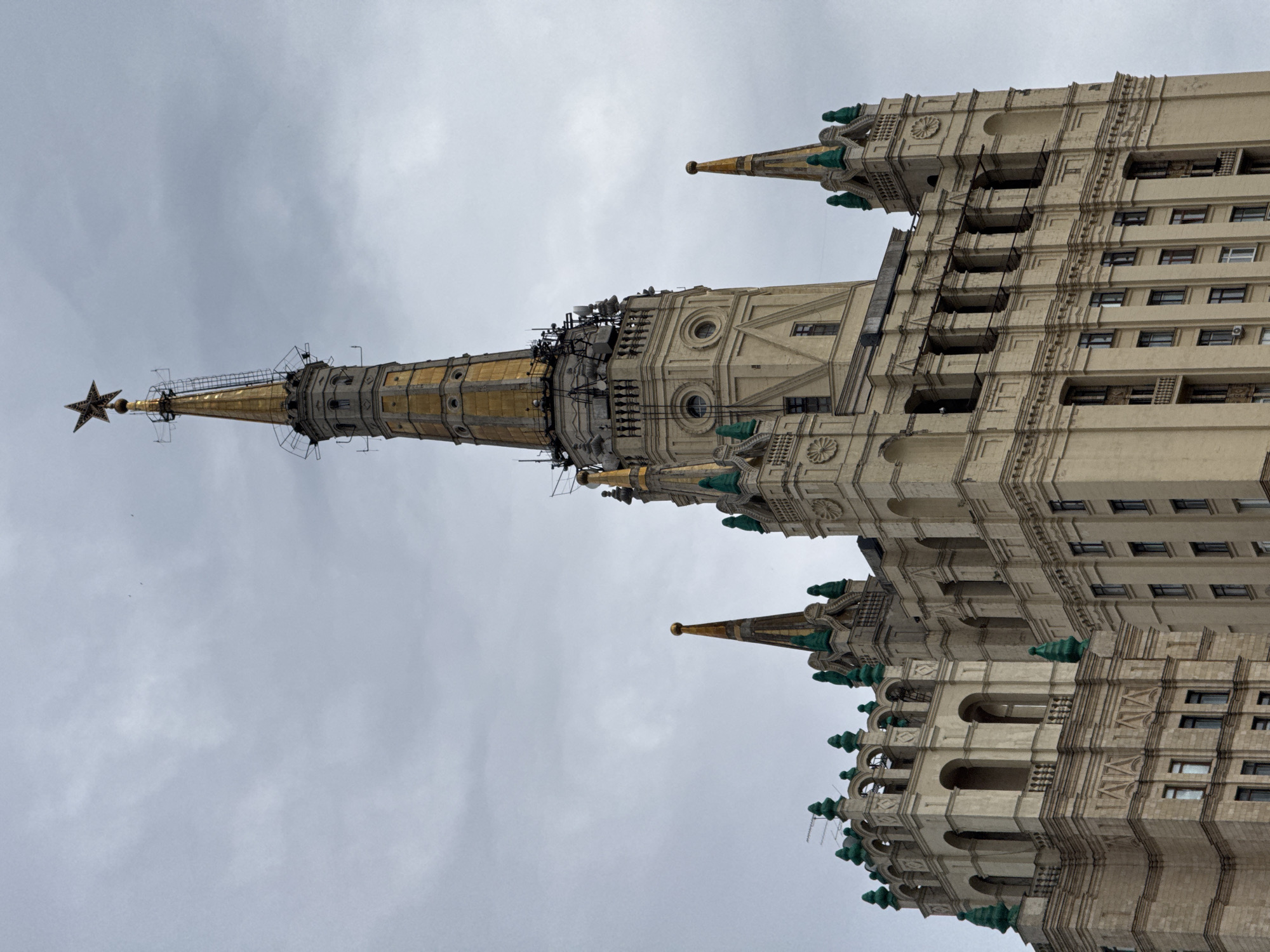 The image depicts the Kotelnicheskaya Embankment Building, one of the "Seven Sisters" skyscrapers in Moscow, Russia. The photograph is taken from a low angle, emphasizing the building's upward reach. The architecture is Stalinist Gothic, characterized by its grand scale, intricate ornamentation, and a central spire topped with a star. The building's facade is light-colored with visible architectural details such as windows, balconies, and decorative elements. The sky is overcast, casting a soft light on the building.