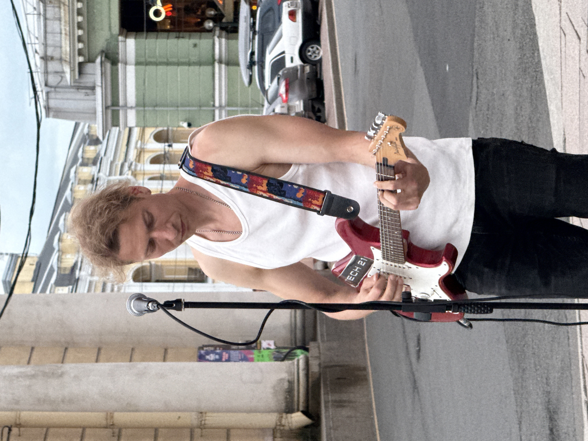 A man with blond hair, wearing a white tank top and black pants, stands on a paved street, playing a red electric guitar. A microphone on a stand is positioned in front of him. In the background, there are classical style buildings with columns, a green building with a neon sign, and several cars parked on the street. The sky is overcast. The scene is likely on Nevsky Prospekt in Saint Petersburg, Russia, given the architectural style and common street performance locations.