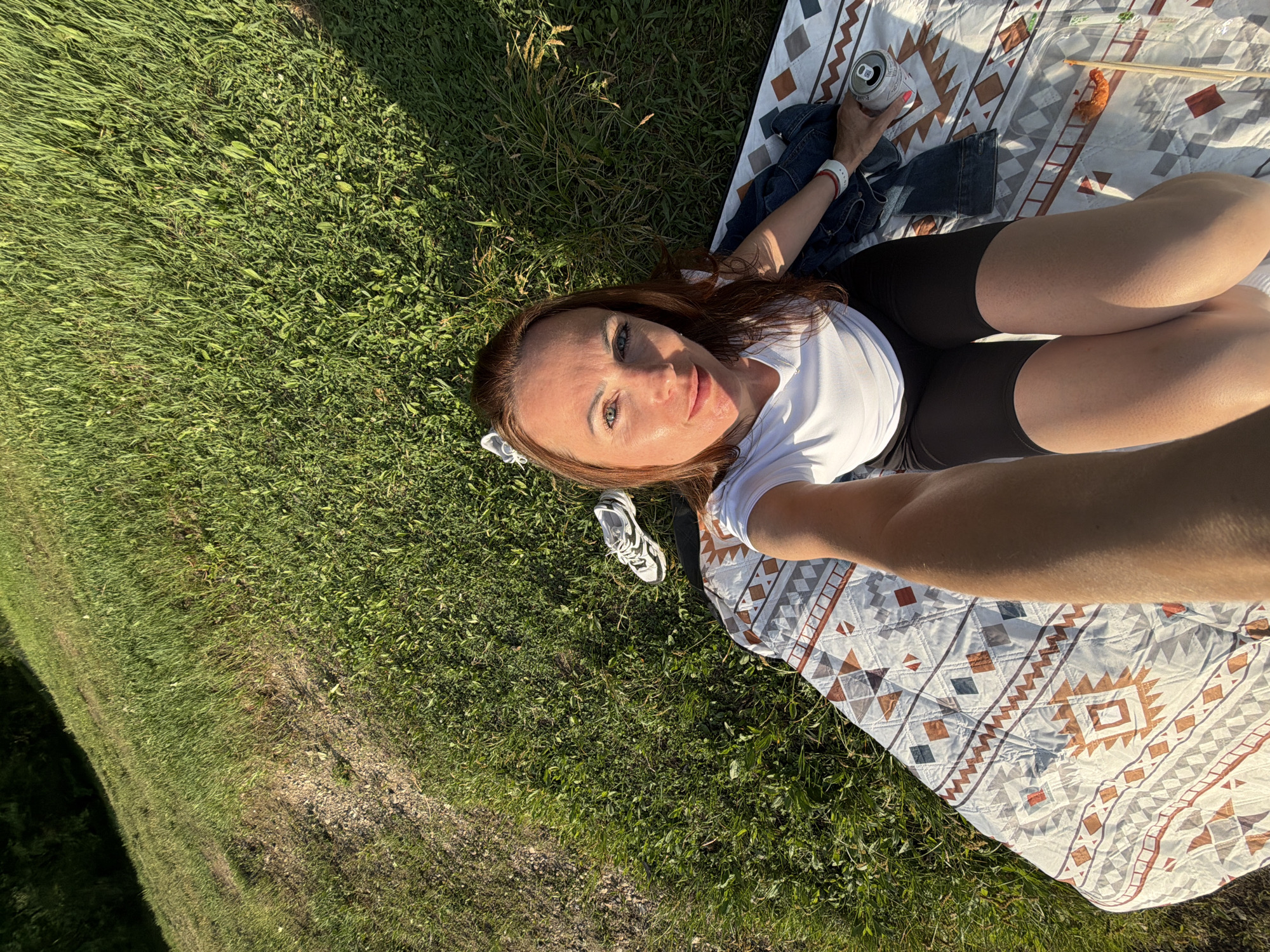A woman is taking a selfie while lying on a patterned blanket on a grassy area. She is wearing a white t-shirt and black shorts. Her left arm is extended towards the camera. A white sneaker is visible on the grass to her upper left. A denim jacket and a can of soda are on the blanket to her right. A small container of food with chopsticks is also on the blanket. The background consists of green grass and some trees in the distance. The sunlight suggests it is daytime. The location is identified as Birobidzhan, Russia.