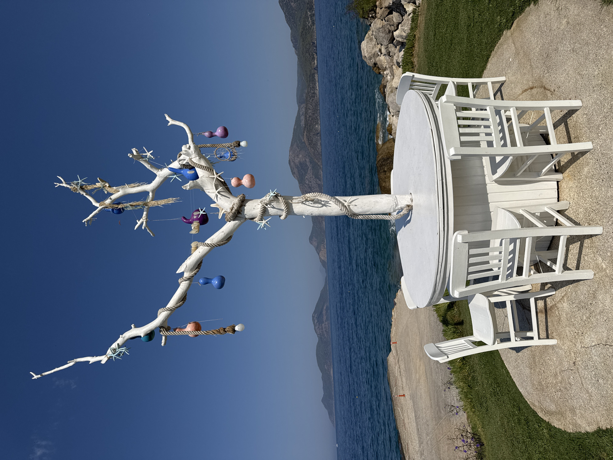 A white, decorative tree sculpture stands behind a white round table with four matching chairs, all set on a concrete patio in Fethiye, Turkey. The sculpture is adorned with colorful gourd-like ornaments, starfish, a dreamcatcher, and rope detailing. The table and chairs are painted white and are arranged around the base of the sculpture. Beyond the patio, a grassy slope slopes down towards a rocky shoreline. The sea stretches out to the horizon, with distant, hilly islands visible. The sky is a clear, bright blue. The scene appears to be an outdoor seating area at a coastal establishment.