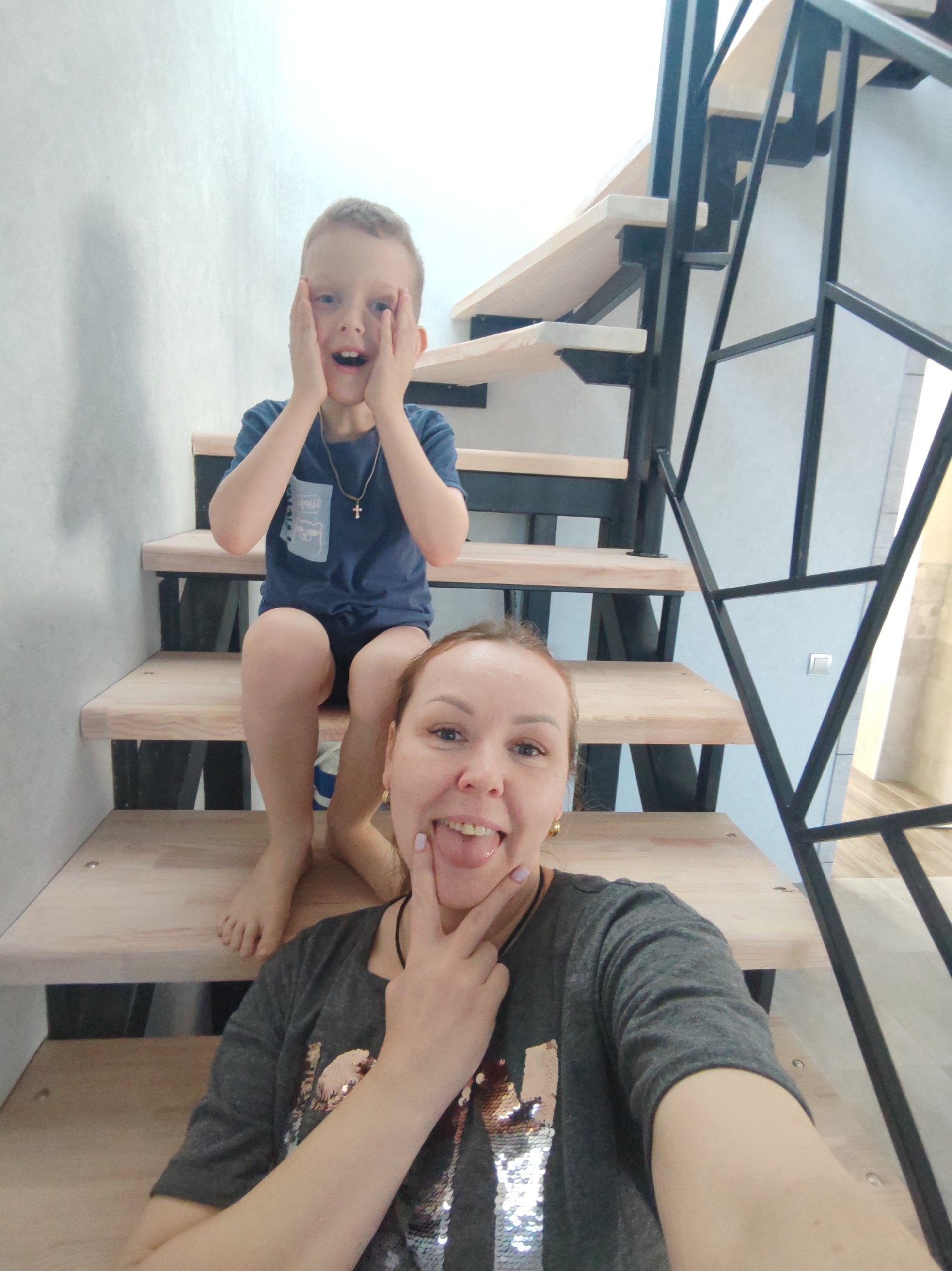 A woman and a boy are posing for a selfie on a staircase. The boy is sitting on a higher step with his hands on his face, looking surprised. He is wearing a blue t-shirt and shorts, and a necklace with a cross. The woman is on a lower step, sticking her tongue out and making a peace sign with her fingers. She is wearing a gray t-shirt. The staircase has wooden steps and a black metal railing. The background is a plain wall. The photo was taken indoors, likely in a home in Chaikovsky, Russia.