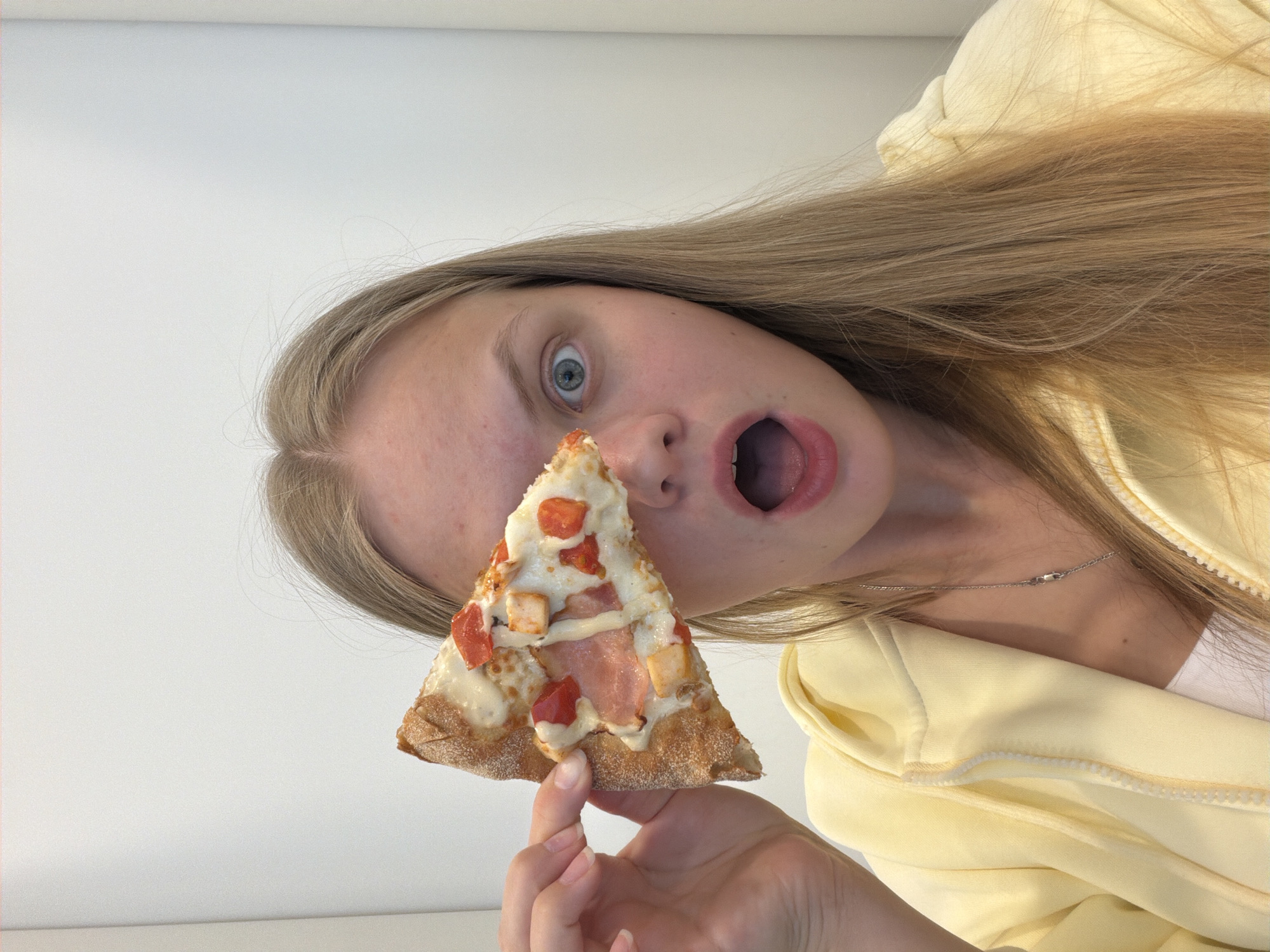 A person with long blonde hair is holding a slice of pizza in front of their face. The person's mouth is open in a surprised or excited expression, and their eyes are wide. They are wearing a light yellow zippered jacket. The pizza slice has a golden crust and toppings that appear to include cheese, tomatoes, chicken, and ham. The background is a plain white wall. The image does not contain any elements that would indicate the location is Izhevsk, Russia.