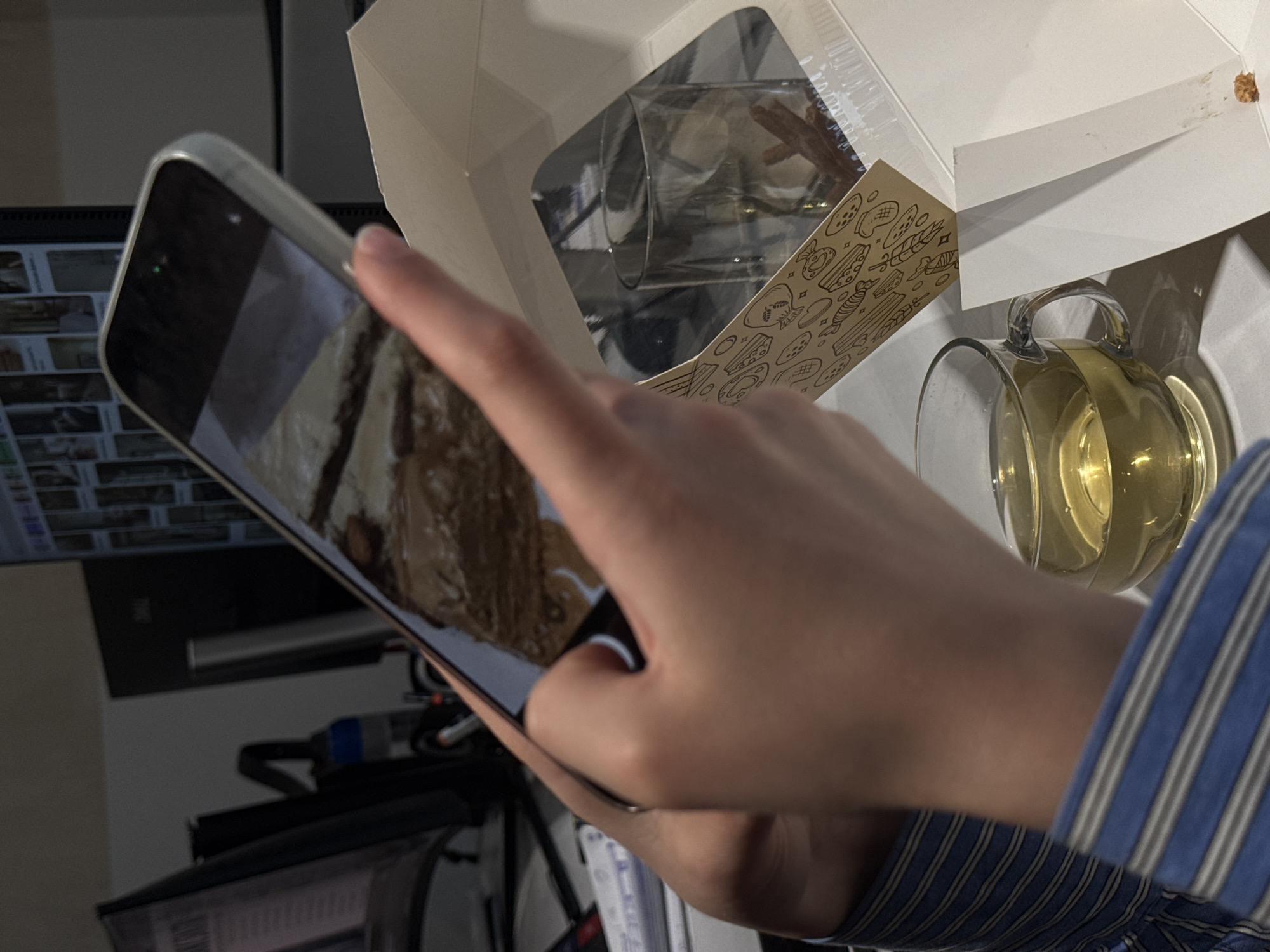 A person holds a smartphone, presumably taking a photograph of a clear glass mug filled with a yellow liquid. The mug is positioned on a white surface next to an open white box with food-related illustrations on its side. The environment includes a computer monitor displaying what appears to be thumbnails of images, along with various wires and equipment. The location of Yerevan, Armenia is not inferable from the image.