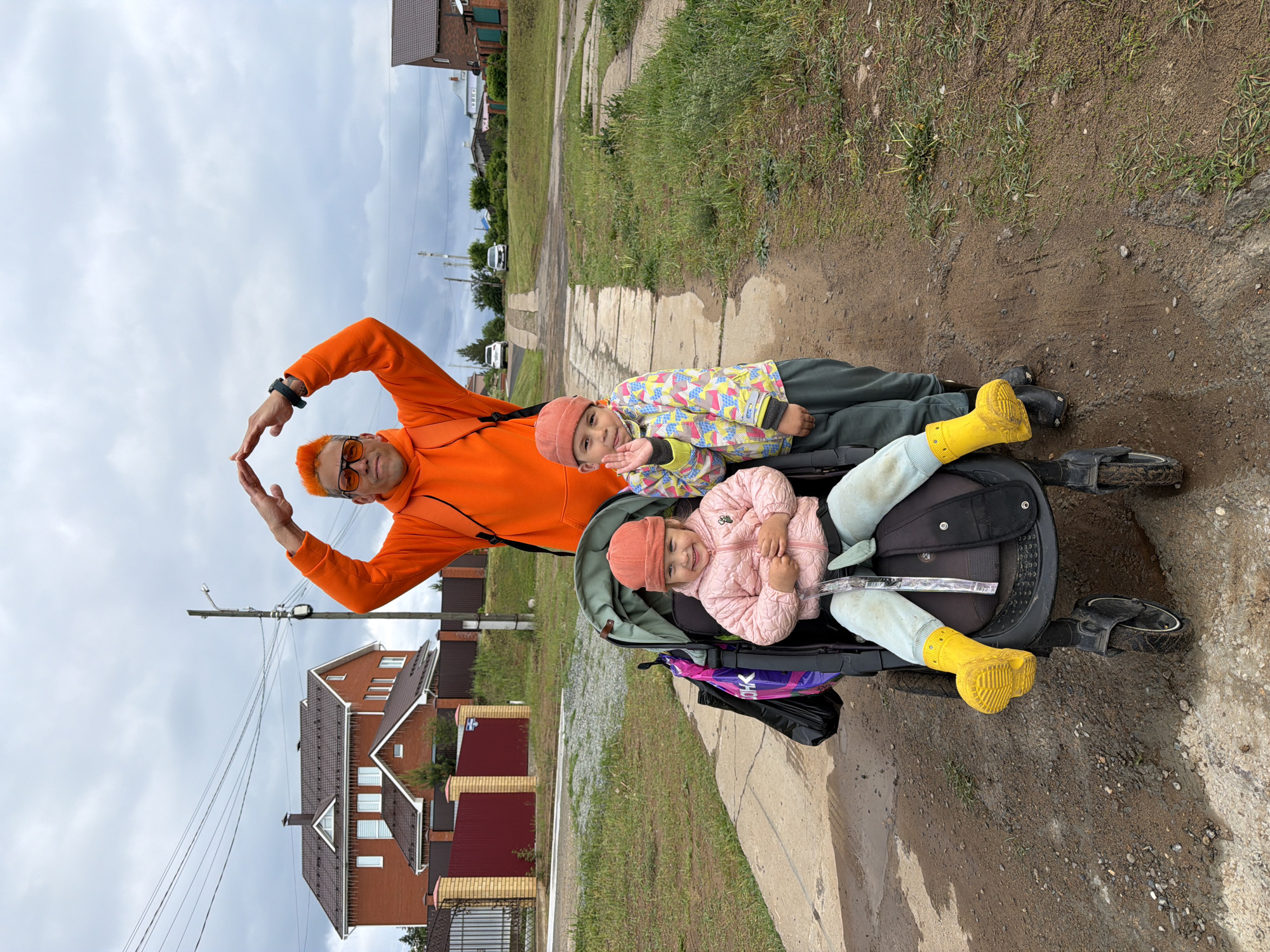 The image captures a father and his two children in the city of Chaikovsky, Russia. The location appears to be a residential street. The father, sporting orange hair, an orange hoodie, and sunglasses, stands behind the stroller, making a heart shape with his hands. One child is seated in a stroller with a pink coat, light blue pants and yellow boots, while the other child lies behind the stroller. The background includes residential buildings, a paved walkway, and a dirt path. The sky is overcast.