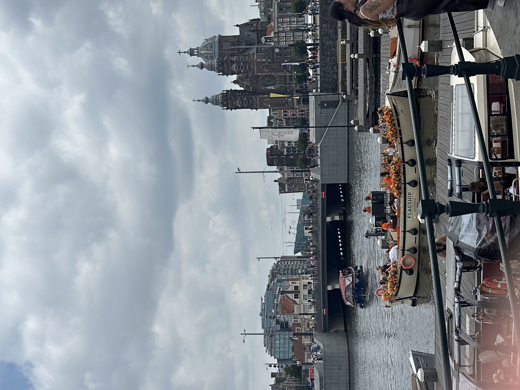 This is a daytime, eye-level, medium shot of a canal in Amsterdam, Netherlands. In the foreground, multiple boats are docked, with a few people visible inside them. The boat closest to the viewer has a clear canopy. To the right, a woman with dark hair and tattoos on her arm stands looking to the right, wearing a white and blue floral top and dark pants. Further into the canal, a white boat labeled "FLAGSHIP AMSTERDAM" is visible, adorned with orange flowers and carrying passengers. Another small red boat is moving away from the viewer on the canal. In the background, a large bridge spans the canal, with numerous people walking across it. Behind the bridge, modern buildings with glass facades are visible, as well as a traditional church with multiple domes and spires. The sky is overcast with clouds.