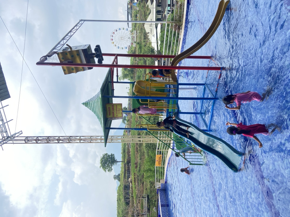 A vibrant scene at a water park. Children are enjoying a playground with slides built over a swimming pool. In the background, a Ferris wheel stands out against the sky. Lush green landscaping surrounds the pool area. The water is a clear blue, adding to the inviting atmosphere. The composition captures the essence of a fun-filled day at the water park.