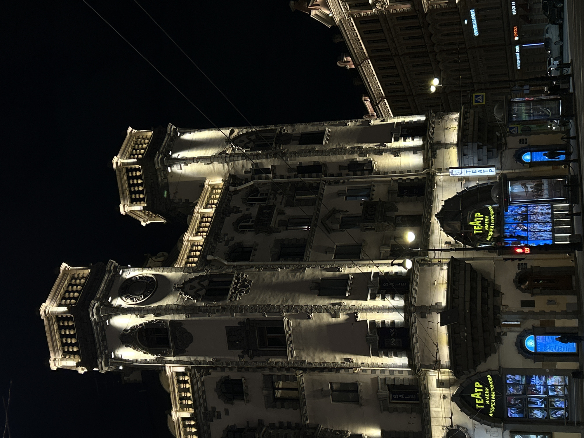 The image, rotated ninety degrees clockwise, captures a brightly illuminated, ornate building at night in an urban setting. The multi-story structure features intricate architectural details, including decorative window frames, cornices, and distinctive tower-like projections at its corners, possibly topped with balustrades. Uplighting and downlighting dramatically highlight the facade's features against the dark sky, where thin overhead wires are visible.
At street level, several establishments are indicated by illuminated signs. A prominent sign in Cyrillic reads "ТЕАТР ИМЕНИ АНДРЕЯ МИРОНОВА" (Theater named after Andrei Mironov), identifying a cultural venue. Other commercial signage includes "COWORKINGSPB.RU," "ОРТОПЕДИЧЕСКИЙ САЛОН" (Orthopedic Salon), "OPTEZA," and "KETCH UP." Multiple "SALE" signs are also visible in window displays. A yellow pedestrian crossing sign with a black "A" is seen further down the street, near what appears to be a street light and some parked vehicles.
The scene conveys a clear night in a city, with artificial lights providing the sole illumination. No people are explicitly visible. The architectural style, prominent Cyrillic text, and the name of the theater strongly suggest the location is in Saint Petersburg, Russia, likely showcasing a historic building like the Dom Knigi (Singer House) or a similar landmark given its distinct towers and central urban location. The overall impression is one of a bustling city center at night, highlighting grand architecture and commercial activity.