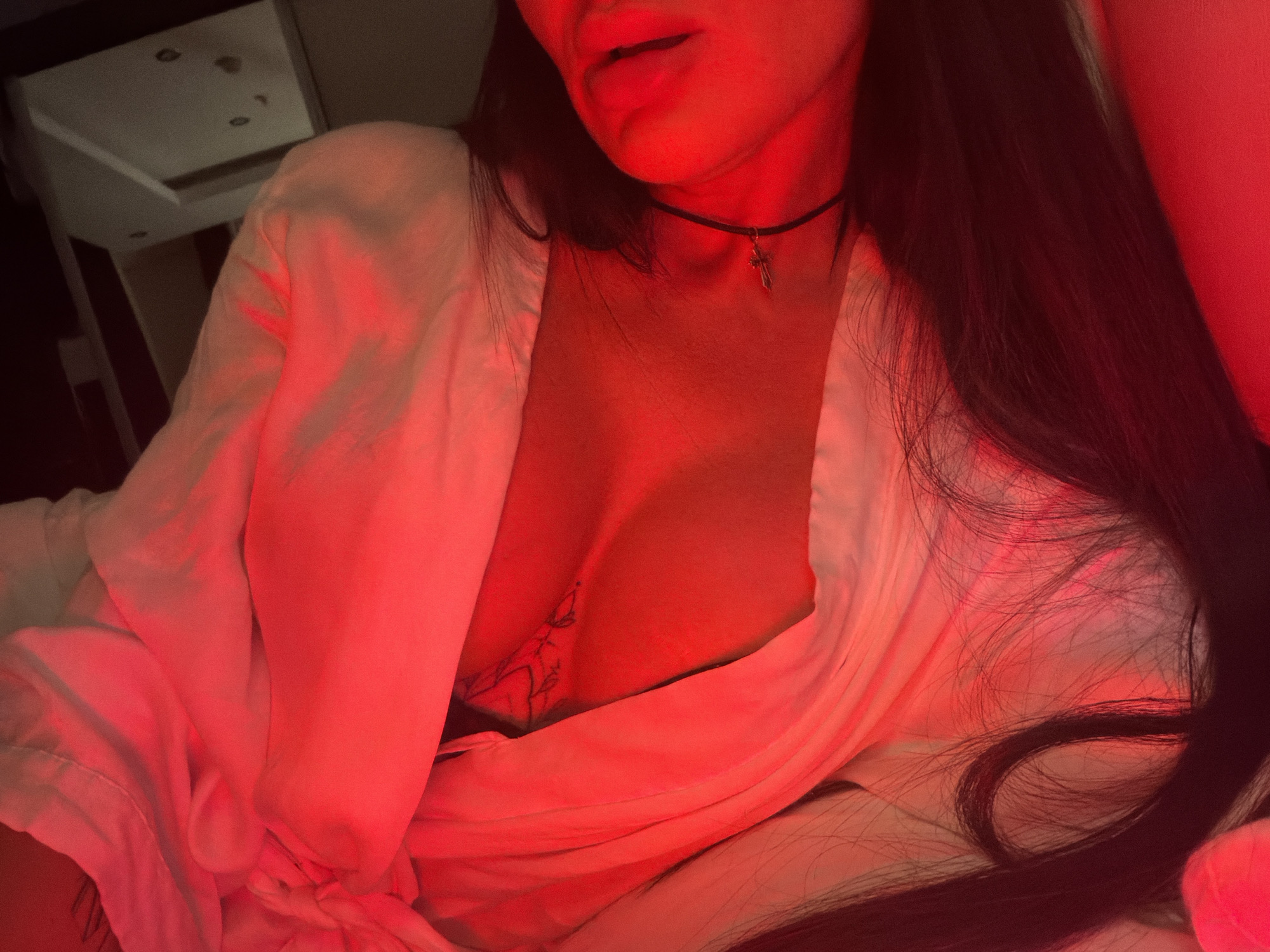 A close-up photograph shows a person partially visible in a red-toned lighting environment. The visible portion of the person includes their mouth, neck, and chest area. The person is wearing a light-colored robe or shirt that is open, revealing part of their chest where a tattoo is visible. They are also wearing a thin black choker with a small cross pendant. The person has long, dark hair that flows over their shoulder. The background includes a ceiling with recessed lighting. The setting is unlikely to be Boreti, Montenegro as there is no evidence of a specific location.