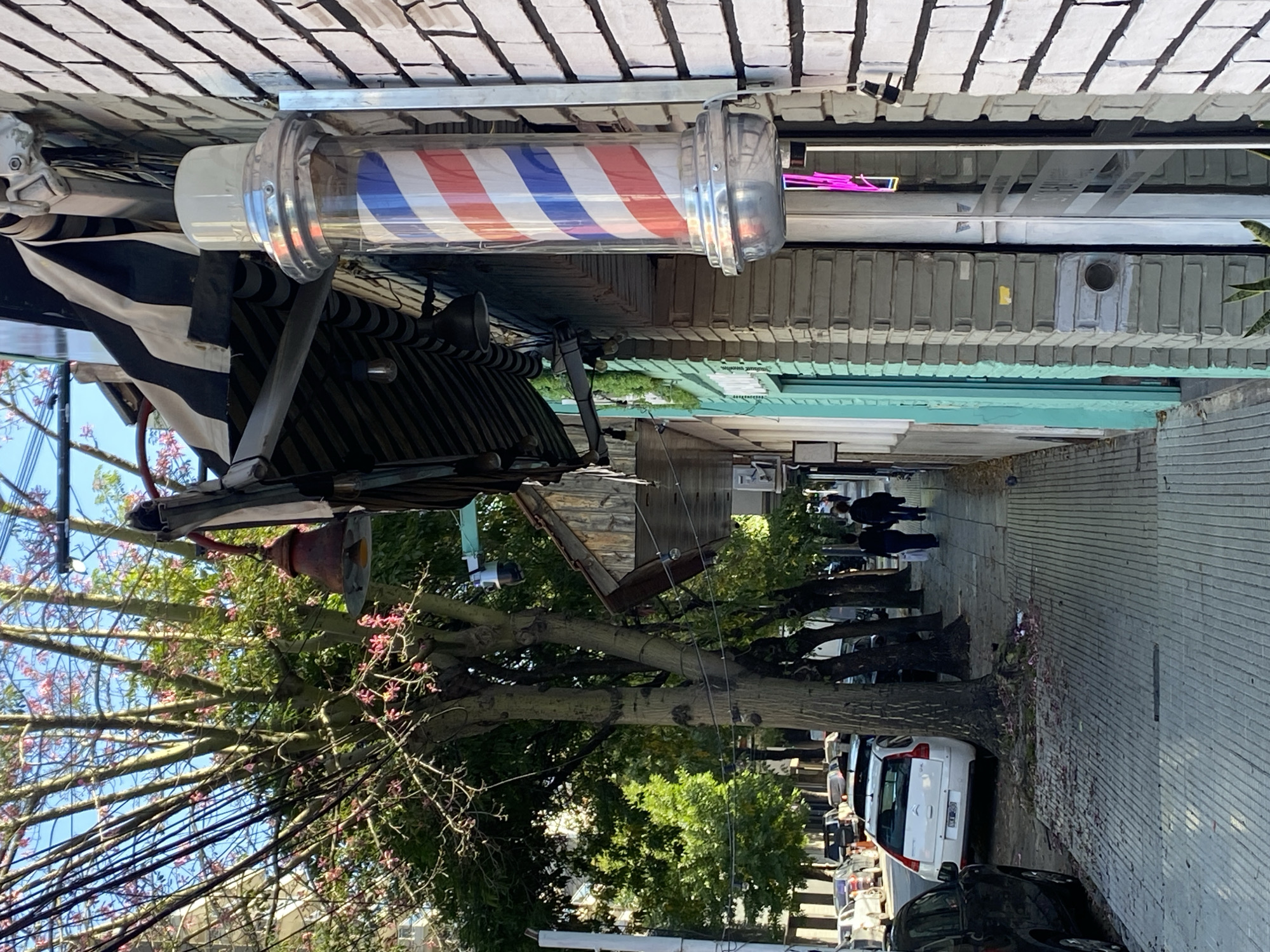The image shows a street view in Buenos Aires, Argentina, with a traditional barber pole mounted on a building. The pole features red, white, and blue stripes encased in a clear cylindrical container with chrome ends. Below the pole, there is a striped awning extending from the building. A tree with blooming pink flowers partially obscures the view. In the background, people are walking along a sidewalk lined with trees and parked cars. The street is paved with what appears to be patterned concrete.