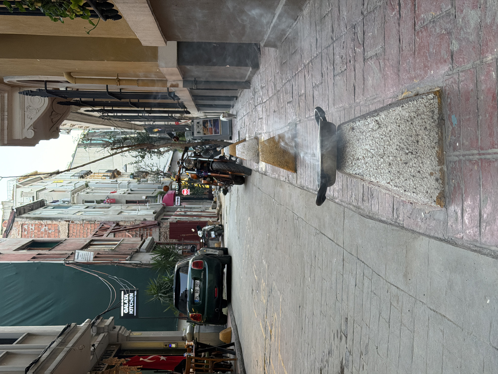 This image captures a narrow urban street or alleyway in Beyoğlu, Turkey, during what appears to be daytime, likely under overcast skies or indirect sunlight. The perspective looks down the street, which is lined with multi-story buildings on both sides.
In the foreground, on the right side of the paved street, a black cat stands poised on a textured concrete planter. A distinct plume of smoke or steam rises from just in front of the cat, creating a point of interest. The street surface transitions from light grey paving blocks in the foreground to a reddish-brown tiled pattern on the sidewalk.
Further down the street, a black motorcycle is parked on the left. Beyond it, a dark green Mini Cooper car is parked, facing away from the viewer. Its rear license plate, "34 BD 987," is clearly visible, indicating its Turkish origin, along with the text "MINI Cooper" on the trunk. A person wearing a pink top is faintly visible walking in the distance near the motorcycle.
The buildings along the street display a mix of architectural styles. On the immediate left, a building features an ornate white corbel and black decorative railings. Further along the left, another building shows exposed brickwork on its upper floors and a large dark green awning or tarp at street level. A prominent white hanging sign attached to this building reads "GALAPA KITCHEN." Above the awning, faint text "TORNACI" can be seen on a building. A partially visible Turkish flag is seen near the bottom left. The overall scene depicts a calm, typical urban street environment with parked vehicles and the distinct presence of a street cat.