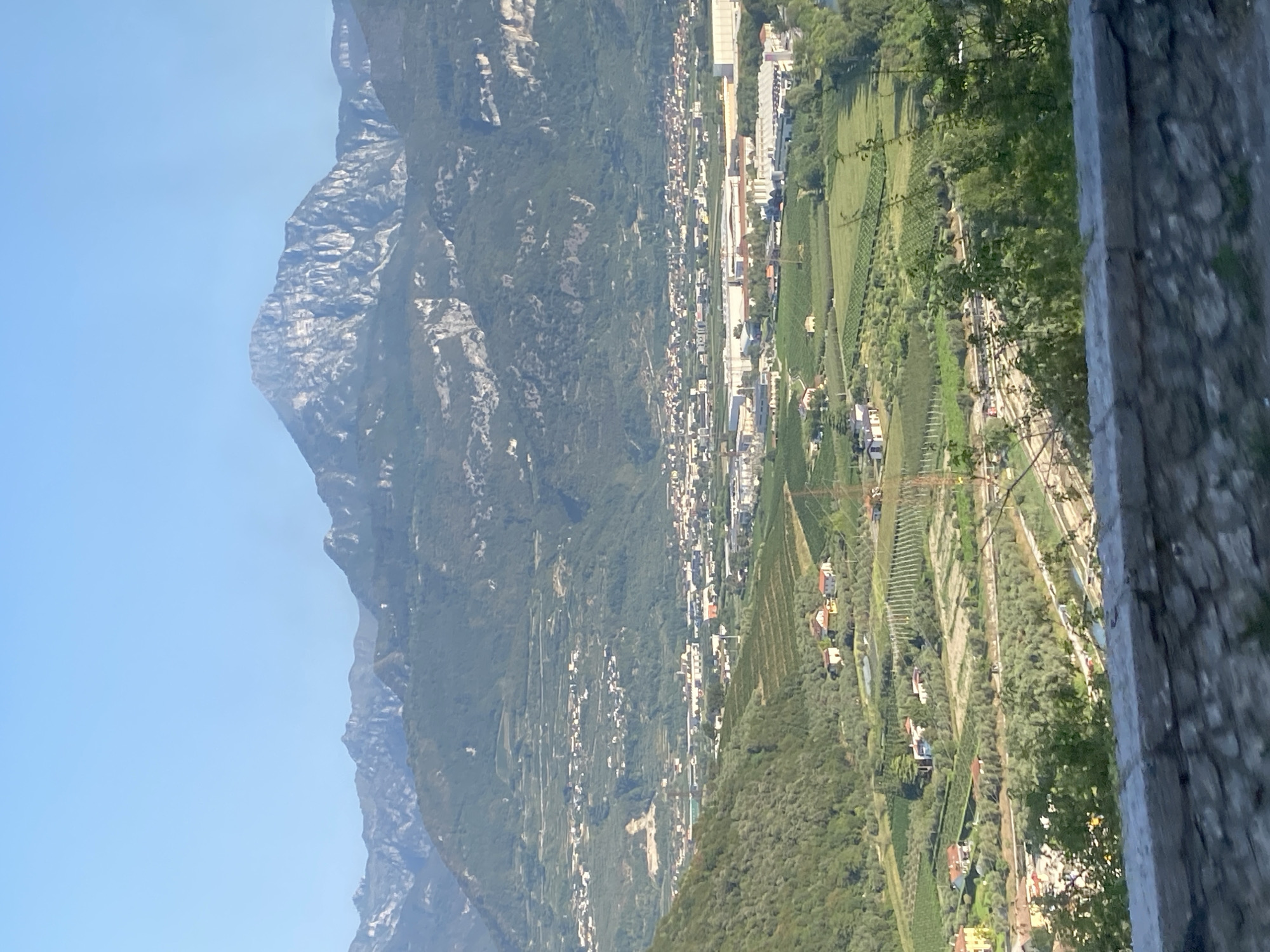 The image shows a landscape view from a high vantage point, looking down upon a valley and a distant mountain range. In the foreground, a stone wall forms a boundary, with a blurred embankment and vegetation below. The middle ground reveals a town or city with a mix of residential buildings and industrial structures, nestled amongst rolling hills covered in vineyards and olive groves. The distant background is dominated by a large, rugged mountain with a distinctly jagged peak, its slopes covered in green vegetation and rocky outcrops. The sky above is clear and blue. The scene appears to be located in Nago, Italy, given the characteristic Italian countryside and mountain scenery. There are no people or animals visible in the image.