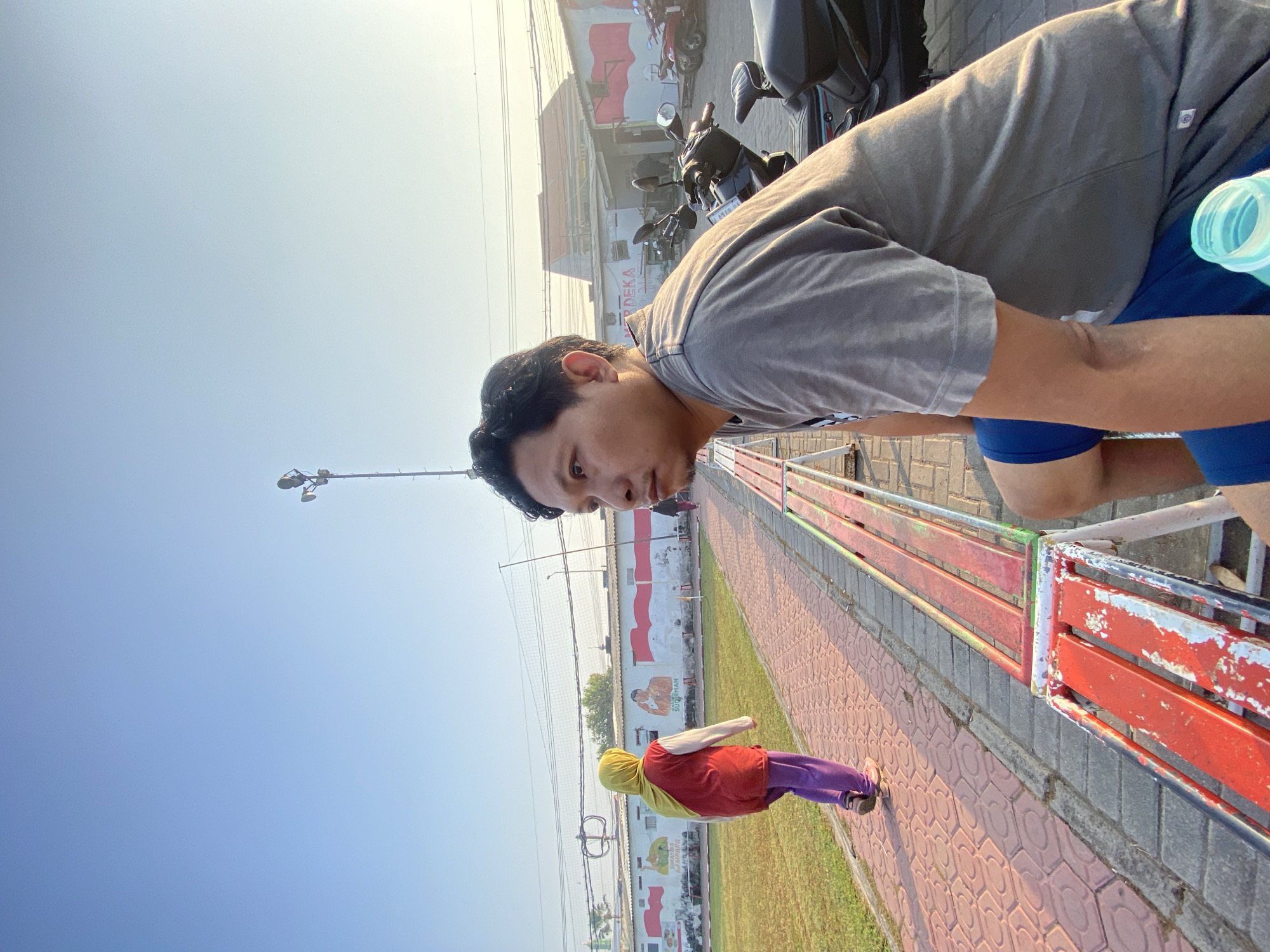 The image depicts an outdoor scene in Sumorame, Indonesia, likely a public area or park, bathed in the soft light of a clear, bright sky. In the foreground, a man wearing a grey t-shirt and blue shorts is seated on a bench, looking to his left. His posture suggests he is resting or observing. A partially visible plastic water bottle is next to him. Behind him, a red and white striped metal bench is prominent. Further back, a person wearing a yellow headscarf and a red and white outfit walks away from the camera along a patterned brick pathway that borders a grassy area. In the background, a low-rise building with a white facade features murals, including depictions of the Indonesian flag and what appear to be historical figures or scenes. A lamppost with multiple lights stands tall against the sky, and several motorcycles are parked near the building. The overall atmosphere is relaxed and daytime.