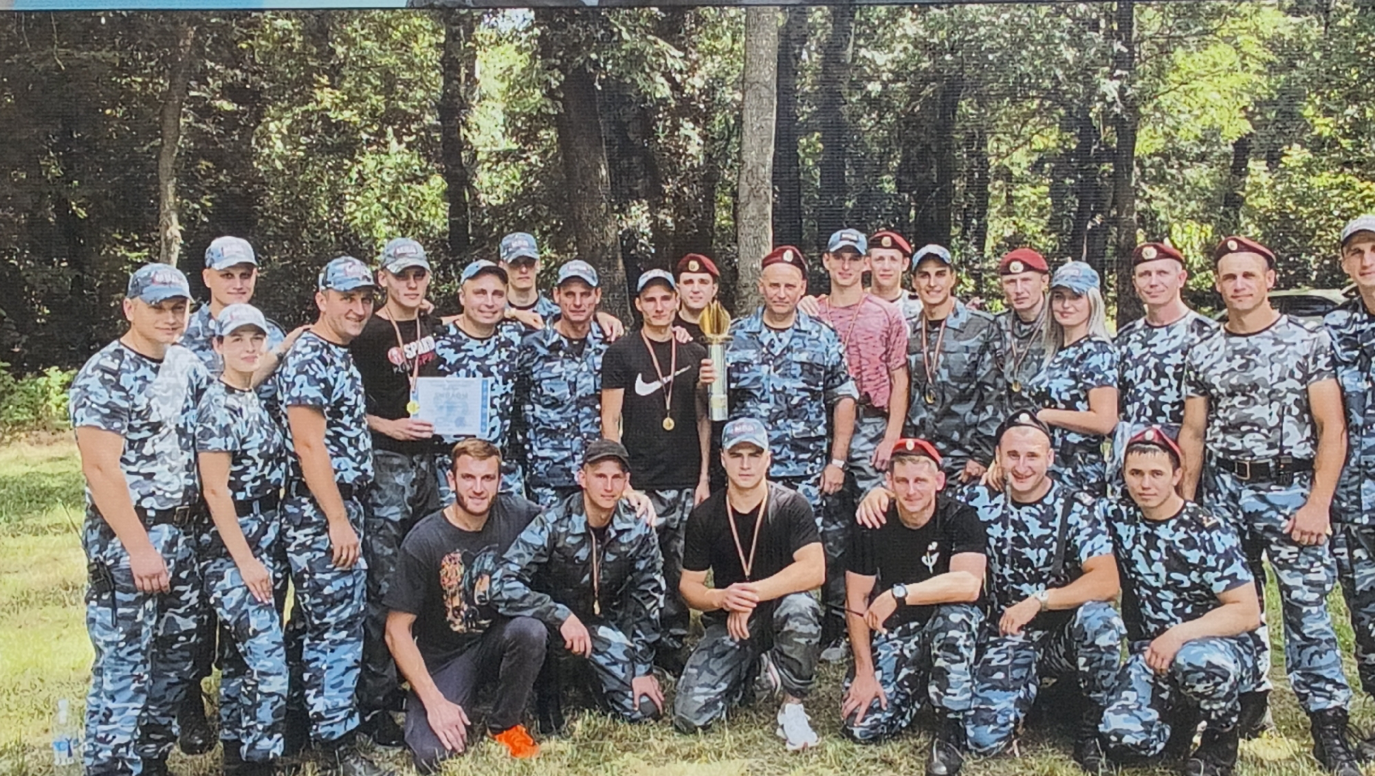 A large group of approximately 25-30 individuals, predominantly male, with a few females, is captured in an outdoor group photograph. The setting appears to be a wooded area or park in Tiraspol, Moldova, with lush green foliage and trees forming the background. The lighting suggests it is daytime, likely a bright morning or afternoon.
The majority of the group is uniformly dressed in blue, grey, and black camouflage t-shirts and pants. Many also wear matching baseball caps, some bearing the Cyrillic letters "МВД" (MVD), likely indicating the Ministry of Internal Affairs. Several men are distinguished by maroon berets. A few individuals wear casual attire, such as black t-shirts (one with a Nike swoosh, another partially showing "SPAR") paired with camouflage or dark pants.
The group is arranged in several rows, with some standing and others kneeling or crouching in the front. The image suggests a post-event gathering, possibly celebrating an achievement or completion of a training exercise. This is evidenced by a gold-colored trophy held by a young man in a black t-shirt, a framed certificate or diploma (partially visible with the Cyrillic word "ДИПЛОМ"), and multiple individuals proudly wearing medals around their necks. Most subjects are smiling or have positive, contented expressions, conveying a sense of camaraderie and accomplishment.