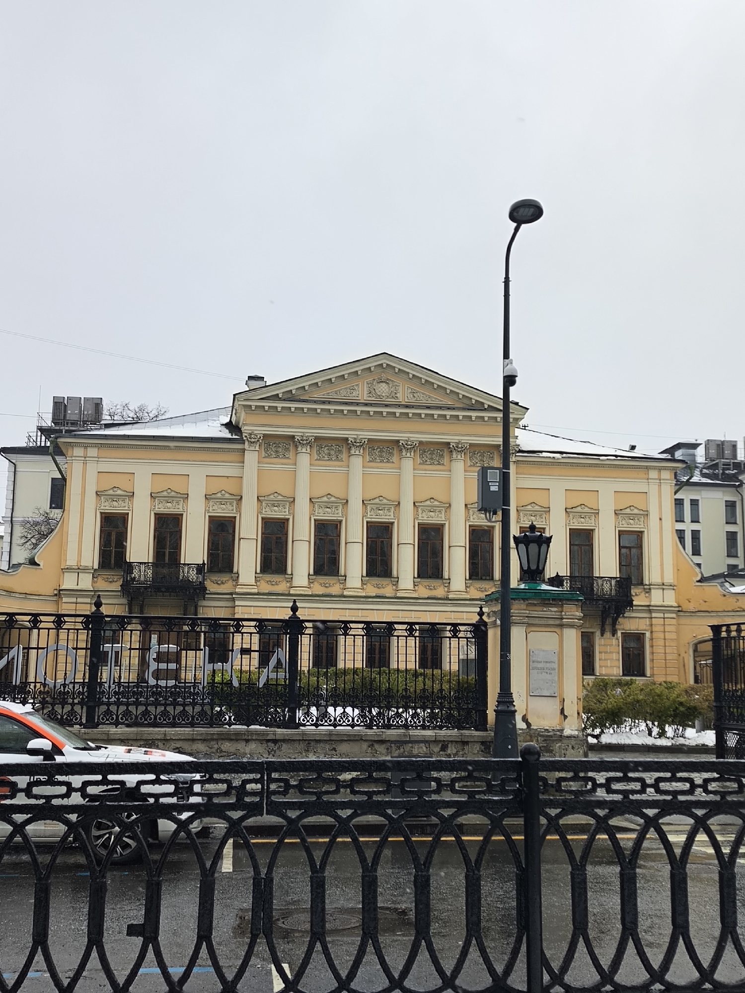 The image presents a grand classical-style building, identified as the Romanov-Sheremetev Palace and housing the Supreme Arbitration Court, located in Moscow, Russia. The building is painted in warm yellow with contrasting white architectural details, including a central portico with classical columns and a decorative pediment. Its facade features numerous tall, dark-framed windows, some accompanied by intricate black wrought-iron balconies. Light snow covers parts of the roof and sparse ground vegetation, indicating a winter or early spring setting.
In the foreground, a multi-layered black wrought-iron fence separates the viewer from the building. The closest fence features an oval-link pattern, while the one behind it displays a more ornate design. Beyond the fences, the street appears wet, suggesting recent precipitation or melting snow. A partial view of a white car with orange stripes is visible on the left side, behind the closer fence.
Two different styles of street lighting are present: a tall, modern black lamp with an oval-shaped head dominates the right side of the frame, while a more traditional, ornate black lantern with a green top rests on a stone pedestal closer to the building. This pedestal bears a plaque with Cyrillic text, clearly identifying the structure as "Высший арбитражный суд" (Supreme Arbitration Court) and "Дворец Романова-Шереметева" (Romanov-Sheremetev Palace). The sky is uniformly overcast and bright grey, contributing to the muted ambiance of the scene. The image captures a quiet, static urban environment with no human activity.