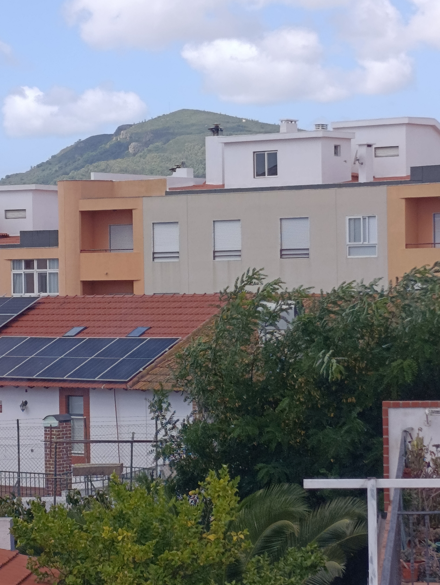 This image depicts a residential area with a view of a hillside in the background. The foreground features rooftops with solar panels and terracotta tiles, along with lush green foliage. The middle ground showcases the facades of several modern buildings, some painted in neutral tones and others in a warm orange hue. These buildings have various windows, balconies, and chimneys. The background is dominated by a verdant, mountainous landscape under a sky with scattered clouds. The scene appears to be a typical residential neighborhood, likely in a warm climate given the vegetation and architecture. The image does not contain any people, actions, or interactions. The location is identified as the city of Setúbal, Portugal.