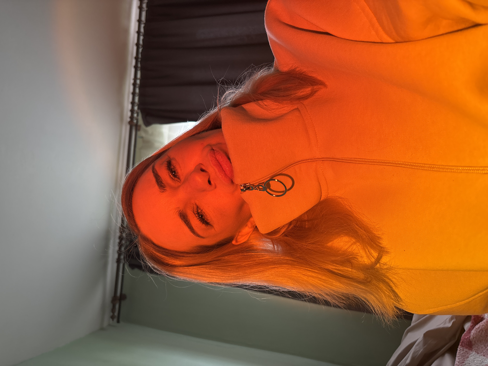 A woman with blond hair is facing the camera, wearing a yellow zippered jacket. The picture is taken indoors with a light source casting an orange hue. A window with dark curtains is visible in the background, along with a light gray wall and part of a bed with bedding.