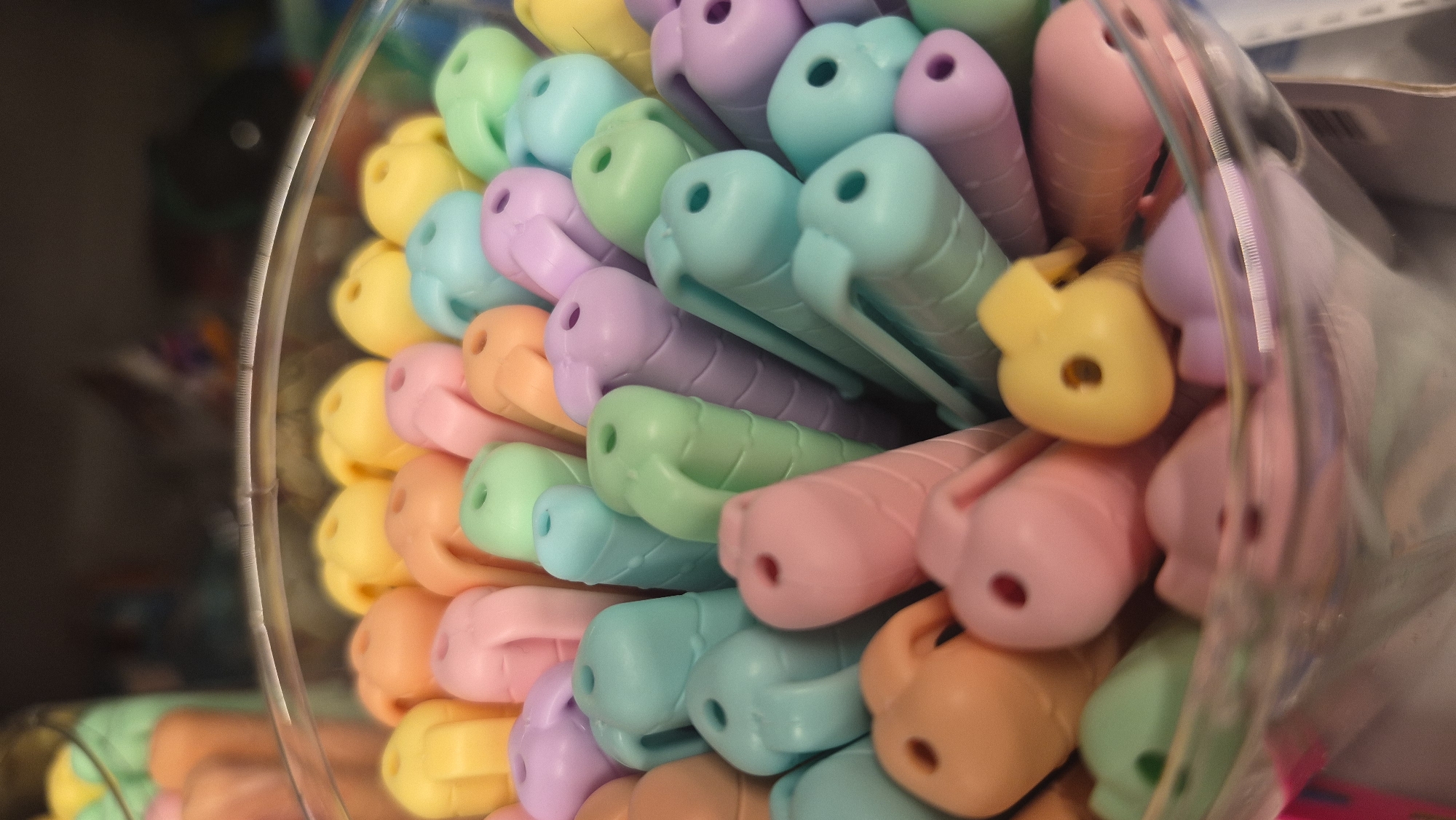 The image is a close-up, shallow depth of field photograph of a collection of brightly colored, plastic Easter-themed candies, likely jelly beans or similar confections, arranged in a clear plastic container. The candies are in pastel shades of yellow, orange, pink, light green, light blue, and lavender. Each candy appears to be molded into a roughly bean-like shape with a small indentation or hole on one end, suggesting they might be intended for stringing or decorative purposes. The container is filled to the brim, and the candies are tightly packed together, creating a textured and vibrant visual. The background is blurred, showing hints of shelves and other merchandise, suggesting this scene takes place in a retail environment. There are no people or animals present. The location in Jēkabpils, Latvia, is not discernible from the visual information provided in the image.