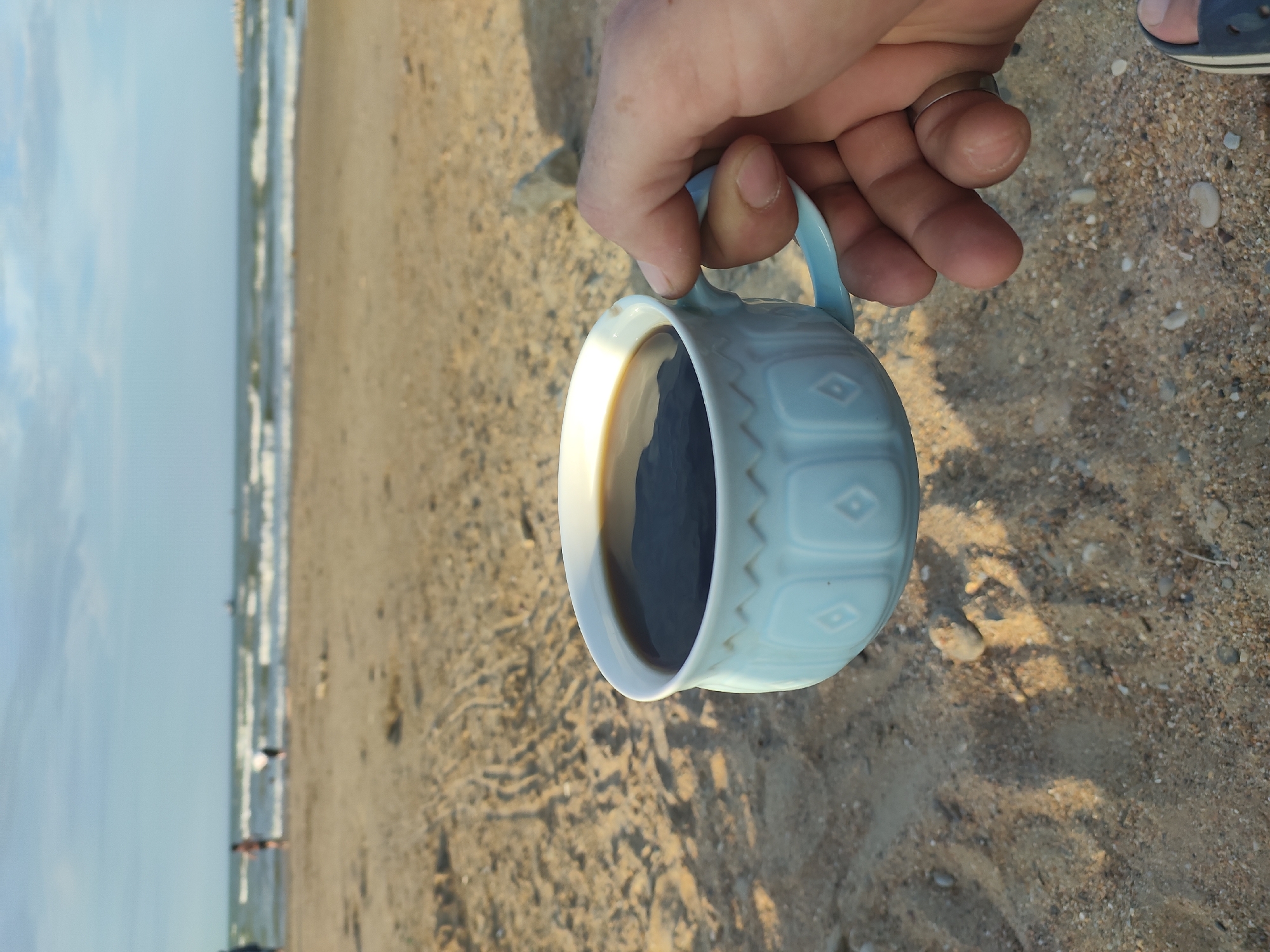 A close-up, eye-level shot shows a person's hand holding a light blue ceramic mug filled with dark coffee. The mug has a decorative pattern of diamond shapes. The mug is positioned on a sandy beach, with the ocean and sky in the background. The sand has tire tracks and small stones. In the distance, there are a few blurred figures walking on the beach and in the water, and a boat on the horizon. The sky is partly cloudy. This scene is consistent with a location on the coast of Makhachkala, Russia, known for its beaches and proximity to the Caspian Sea.