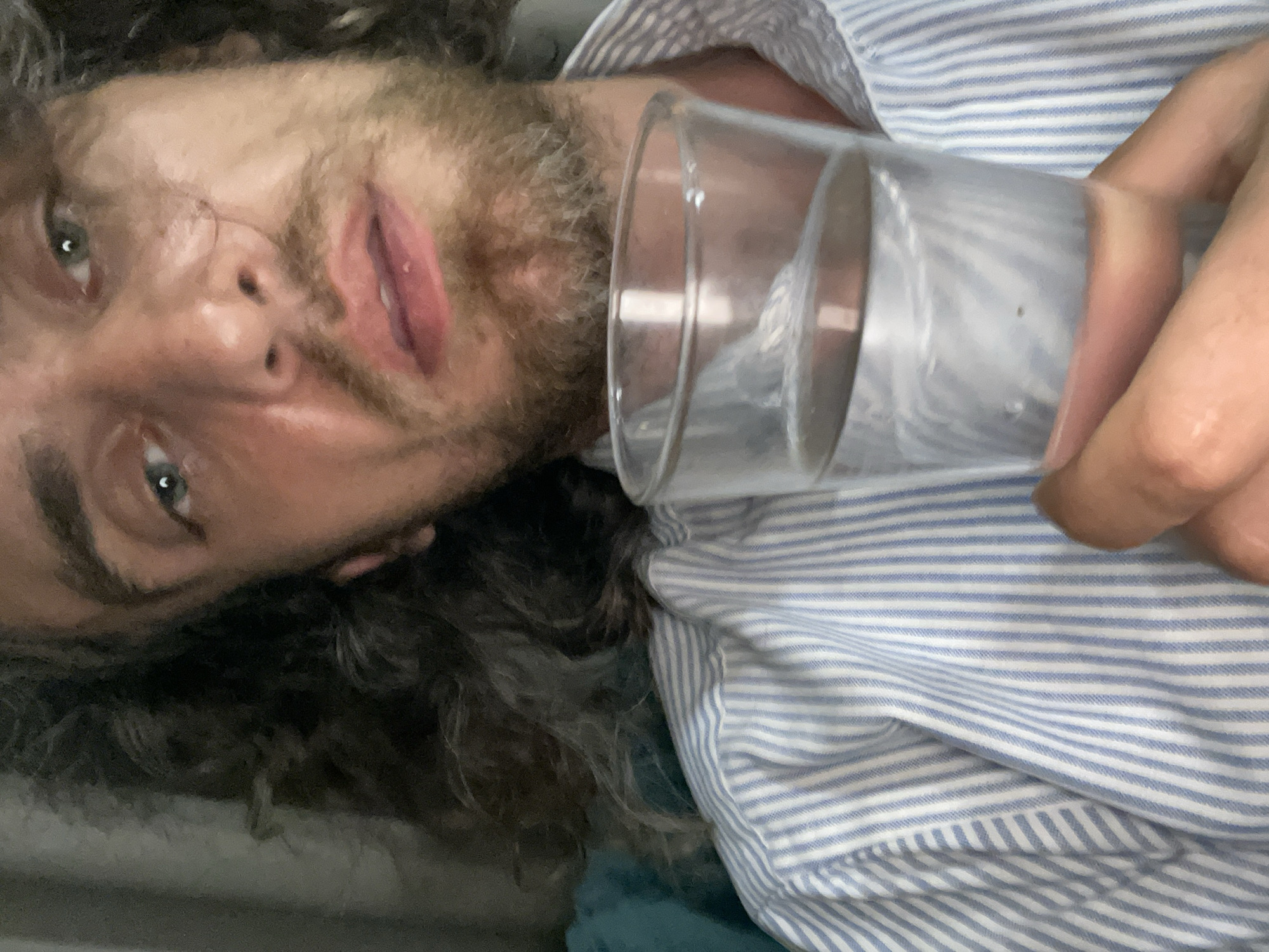A close-up, selfie-style shot shows a fair-skinned person with long, dark, curly hair, wearing a light blue and white striped button-down shirt. The person's face is partially obscured by a glass of water held in their right hand. Their facial features include light-colored eyes, a nose, and lips. The background is out of focus, but appears to be indoors, possibly with a gray curtain on the left. The image is taken in the city of Sakhu, Thailand.