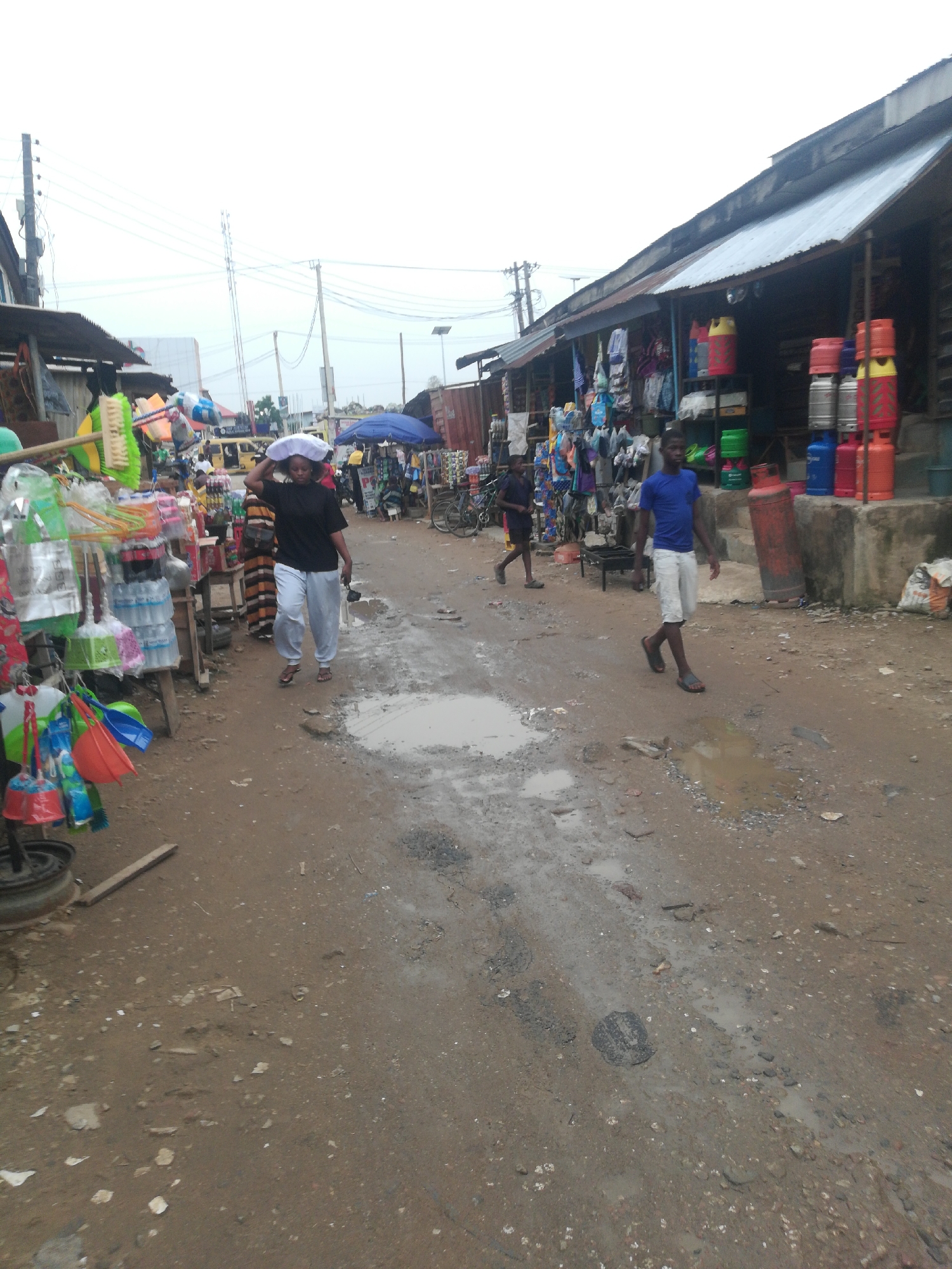 The image captures a street scene in Unknown, Unknown. The ground is uneven and muddy, with puddles of water scattered across the surface. People are walking along the street, and stalls line the sides, displaying various goods for sale. Buildings with corrugated metal roofs are visible in the background. Gas cylinders are stacked outside one of the buildings. Overhead, power lines crisscross the sky.