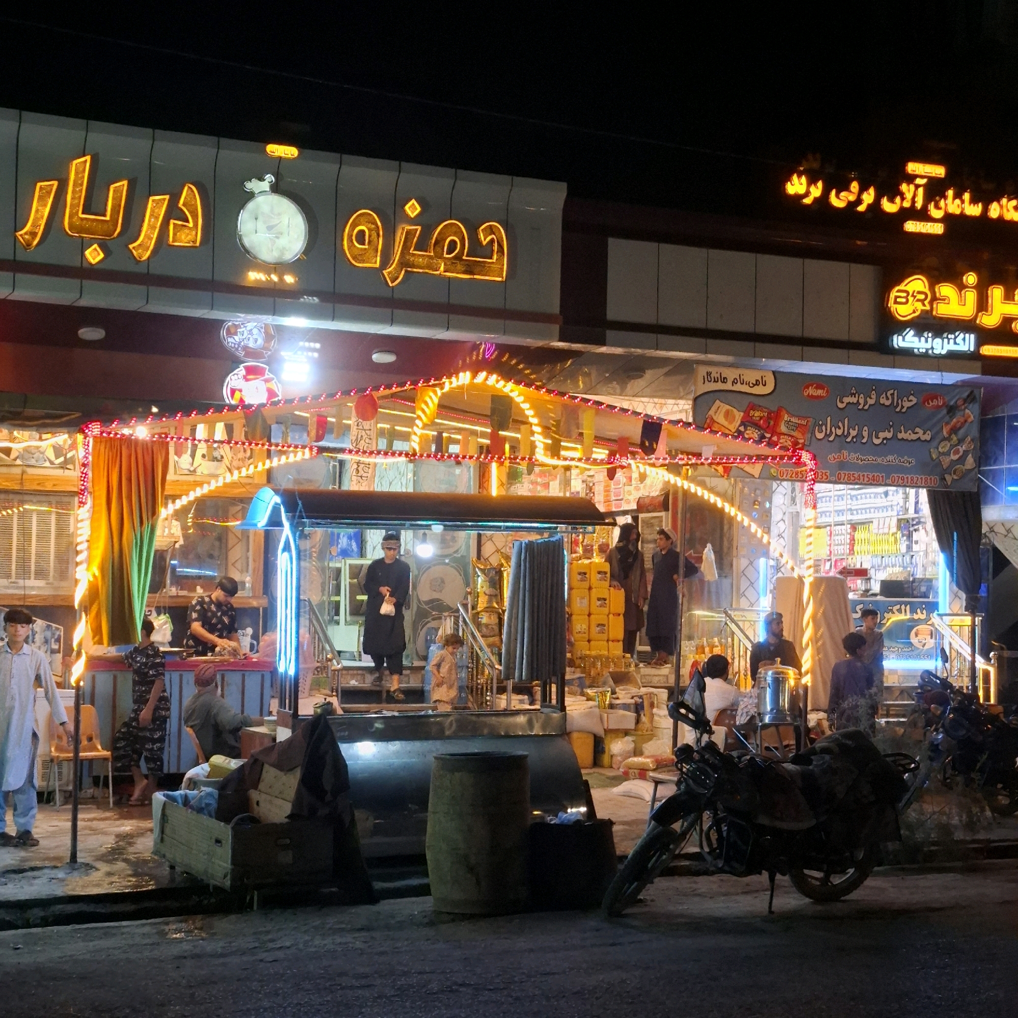 This image captures a nighttime street scene in Sar-e Pol, Afghanistan. A brightly lit storefront is the central focus, adorned with illuminated signs in both Arabic script and English lettering. The storefront appears to be a shop or restaurant, with goods displayed both inside and outside.
Several individuals are visible. In the foreground, a young boy in a light-colored shirt walks towards the left. To the right of him, a man wearing a patterned shirt and a cap is seen preparing something at a counter, possibly food. Further back, other individuals stand or move about near the storefront. A motorcycle is parked prominently in the lower right quadrant of the image, leaning on its kickstand.
The environment suggests a bustling urban setting. The street surface is dark and appears wet in places, reflecting the ambient light. The storefront itself is decorated with strings of colorful lights, enhancing the vibrant atmosphere. The architecture is characteristic of a commercial area, with awnings and shelving visible. The overall impression is one of an active commercial hub, illuminated and busy even after dark.