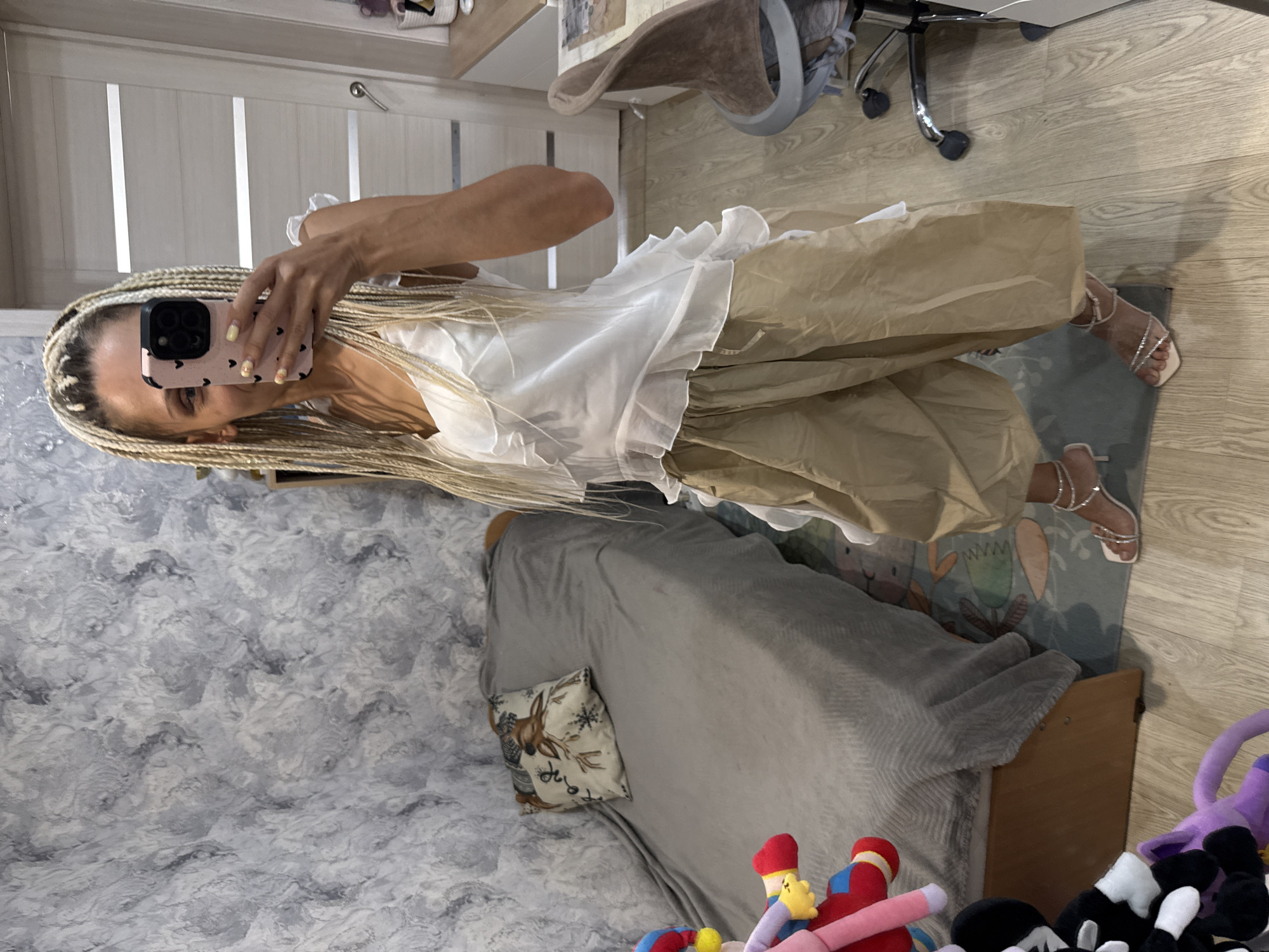 The image shows a woman taking a mirror selfie in a room. She has blonde braids and is wearing a white ruffled top and beige pants. She is also wearing strappy, jeweled sandals. In the background, there is a bed with a grey blanket and a decorative pillow with a deer on it, a wooden dresser, and a wall with a marble-like pattern. To the right of the bed, there are some stuffed animals and a glimpse of a patterned rug. A portion of a desk and a chair are also visible. The lighting suggests it is daytime, possibly indoors as there is no visible outdoor scenery. The overall scene appears to be a casual personal space, likely a bedroom.