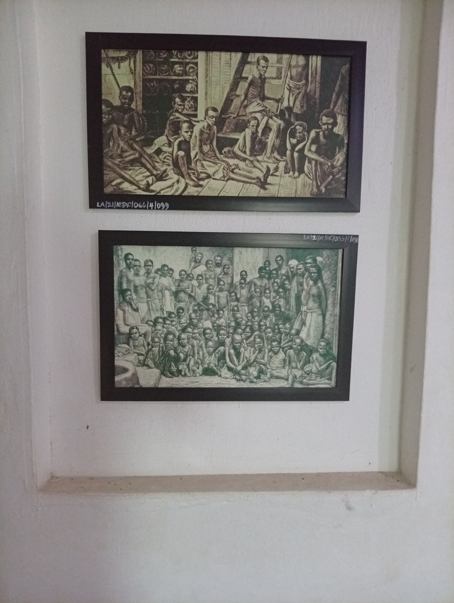 Here's a detailed description of the image:
The image shows two framed pictures hanging on a white wall within a recessed niche. The location is the city of Badagry, Nigeria. Both pictures are in dark brown frames.
The top picture depicts a group of emaciated people, mostly men and boys, in what appears to be a confined space. Some are seated on the floor, while others are on a raised platform or ladder.
The bottom picture portrays a larger group of similarly thin people, including women and children, seated or squatting on the ground. A few individuals stand among them.
Below each picture, a series of numbers and letters is written: "LA/21/MTAC1066/4/099" and "LAT21/NTAC/066/4/0%".
The wall appears to be made of plaster, and the recessed niche has a slightly discolored horizontal ledge.