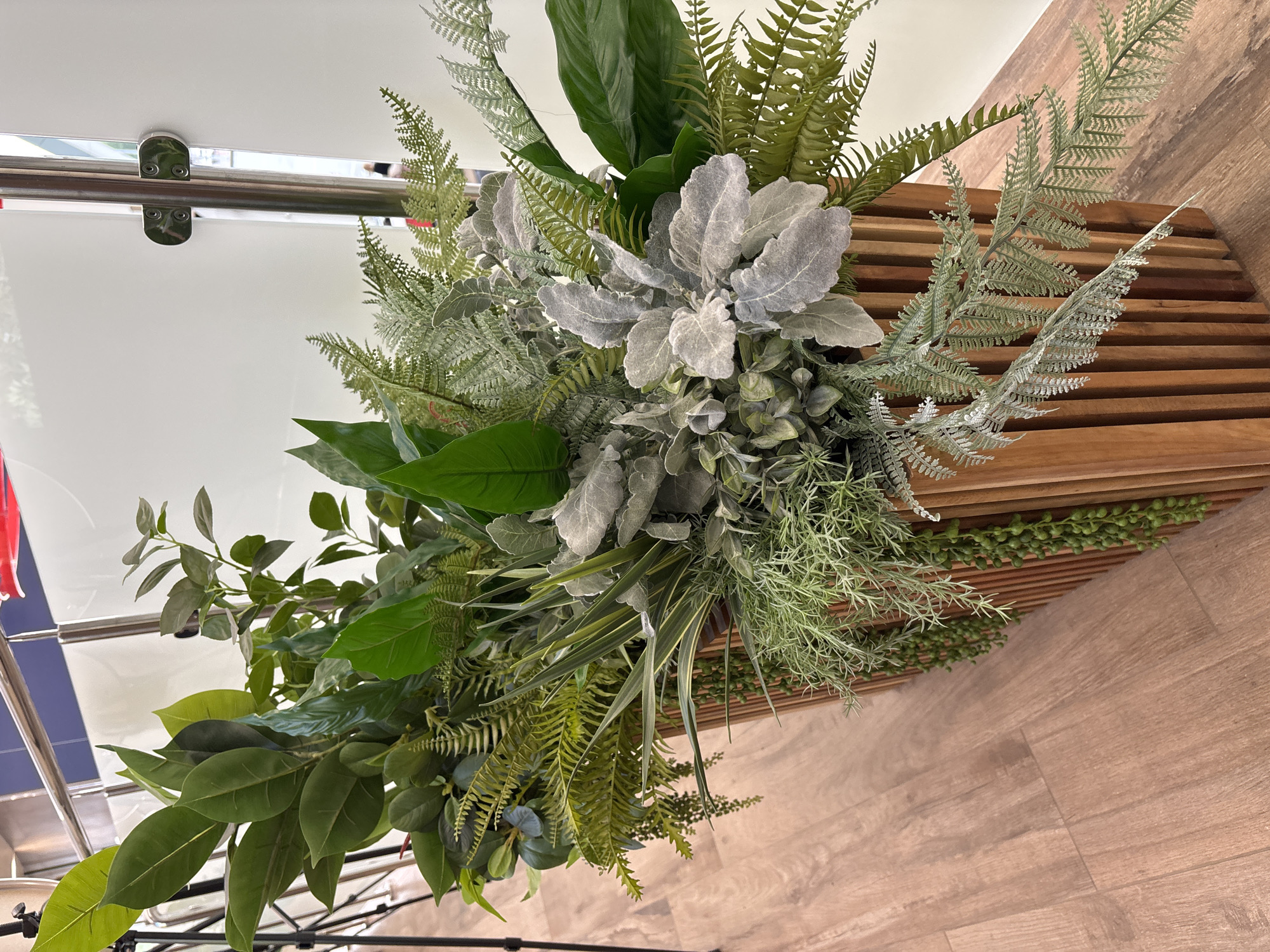 This is a media file of a decorative arrangement of artificial plants. The arrangement features a variety of green foliage, including large, glossy leaves and delicate fern-like fronds. There are also lighter, silvery-gray leaves that add contrast. The plants are densely packed and appear to be arranged in a bouquet or centerpiece. The background shows a light-colored, reflective surface, possibly a wall or a large piece of furniture, with a metallic railing. To the right, there is a wooden surface with horizontal slatted detail, and below that, a wooden floor. The lighting suggests an indoor setting, likely during the day. There is no visible text, people, or any specific activity or event taking place. The setting appears to be a domestic or commercial interior, possibly a lobby or a decorated space. The overall impression is one of artificial greenery used for decoration. The provided location context, Старое Мартьяново, Russia, does not offer any specific cues visible within the image itself.