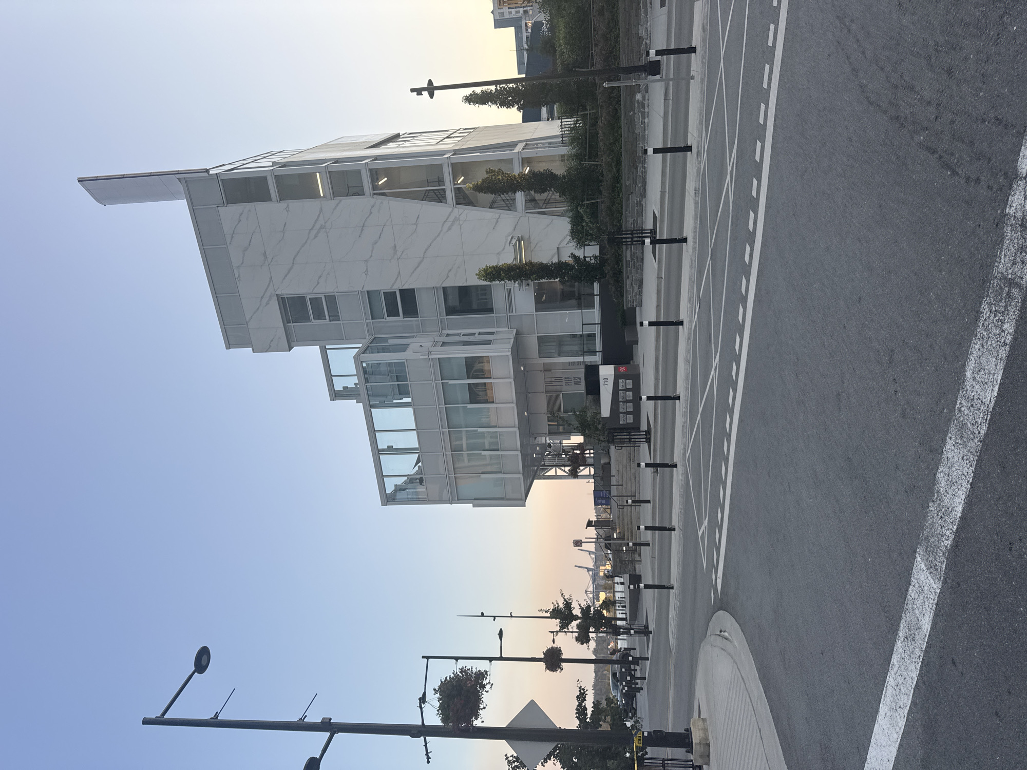 This is a photograph of the Pier West building at 710 Quayside Drive in New Westminster, Canada, taken during the late afternoon or early evening. The building is modern with a white, angular facade, featuring large windows and a distinctive, tall, white vertical element at its peak. The sky is clear with a gentle gradient from pale blue to a peachy hue near the horizon, suggesting sunset.
In the foreground, a paved road with white lane markings curves to the left. Black and white bollards line the sidewalk. To the left of the road, a lamppost with hanging flower baskets stands tall. Beyond the sidewalk and a low stone wall, the Pier West building is visible. Its entrance appears to be at street level, with a set of stairs leading up to a higher level. The building is surrounded by some greenery and trees.
In the distance, across a body of water, industrial structures and cranes are visible, suggesting a port or industrial area. The overall atmosphere is calm and the lighting is soft, characteristic of twilight. There are no visible people in the frame. The image captures a quiet urban waterfront scene.