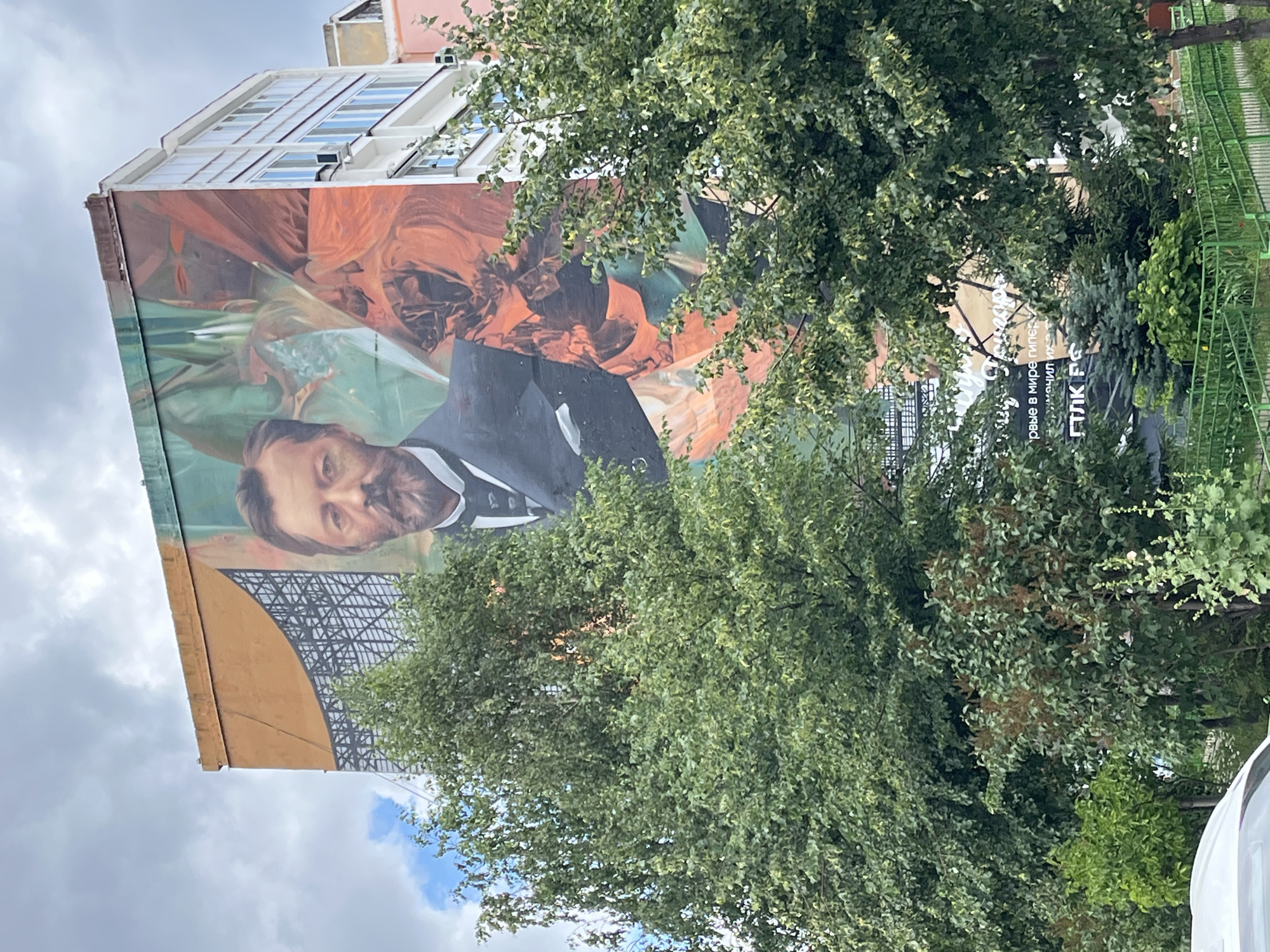 The media file shows a mural painted on the side of a building in Nizhny Novgorod, Russia. The mural depicts a portrait of a man with a beard, wearing a suit. He has a serious expression. The background of the mural features abstract shapes and colors, with hints of orange and green.
The building appears to be an apartment complex with multiple stories. The top of the building has windows with a modern design. Trees with green foliage partially obscure the mural and the building. A green metal fence is visible to the right of the frame, suggesting the mural is located in a public area, possibly a park or residential courtyard.
The sky is cloudy, indicating overcast weather. The lighting suggests it is daytime, but the specific time is unclear due to the cloud cover.
Visible text on a sign in the background is difficult to read clearly due to its distance and partial obstruction by trees. However, some Cyrillic letters are discernible. One section appears to read "РОВЫЕ В МИРЕ ТИПЕР", and another segment might say "ПЛК РС". The context of this text is not clear from the image.
The overall impression is that of an urban art installation in a residential or public space.
