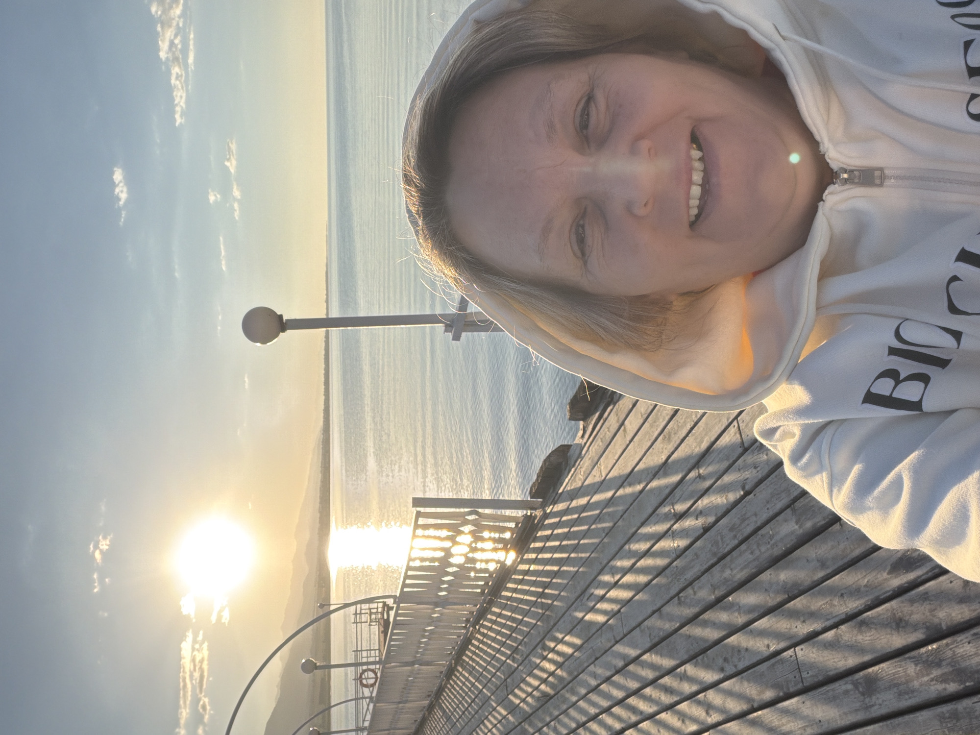 A selfie is taken by a woman wearing a white hooded sweatshirt with "BIO" visible on the front. She is standing on a wooden pier with a white railing. The sun is setting over a body of water, casting long shadows across the pier. In the background, mountains are visible on the horizon. The sky is clear with a few scattered clouds.