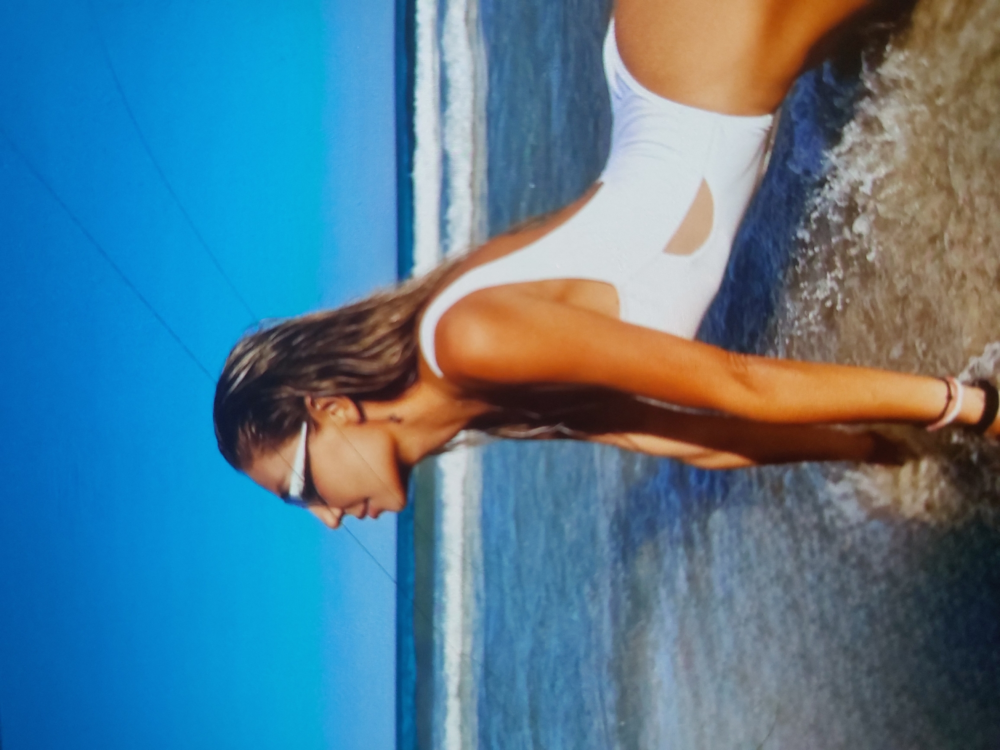 A woman in a white swimsuit is in the water at the beach. The background shows the sea and sky. The location is likely Tsarevo, Bulgaria, based on the prompt's instruction. The woman is tanned and wearing sunglasses.