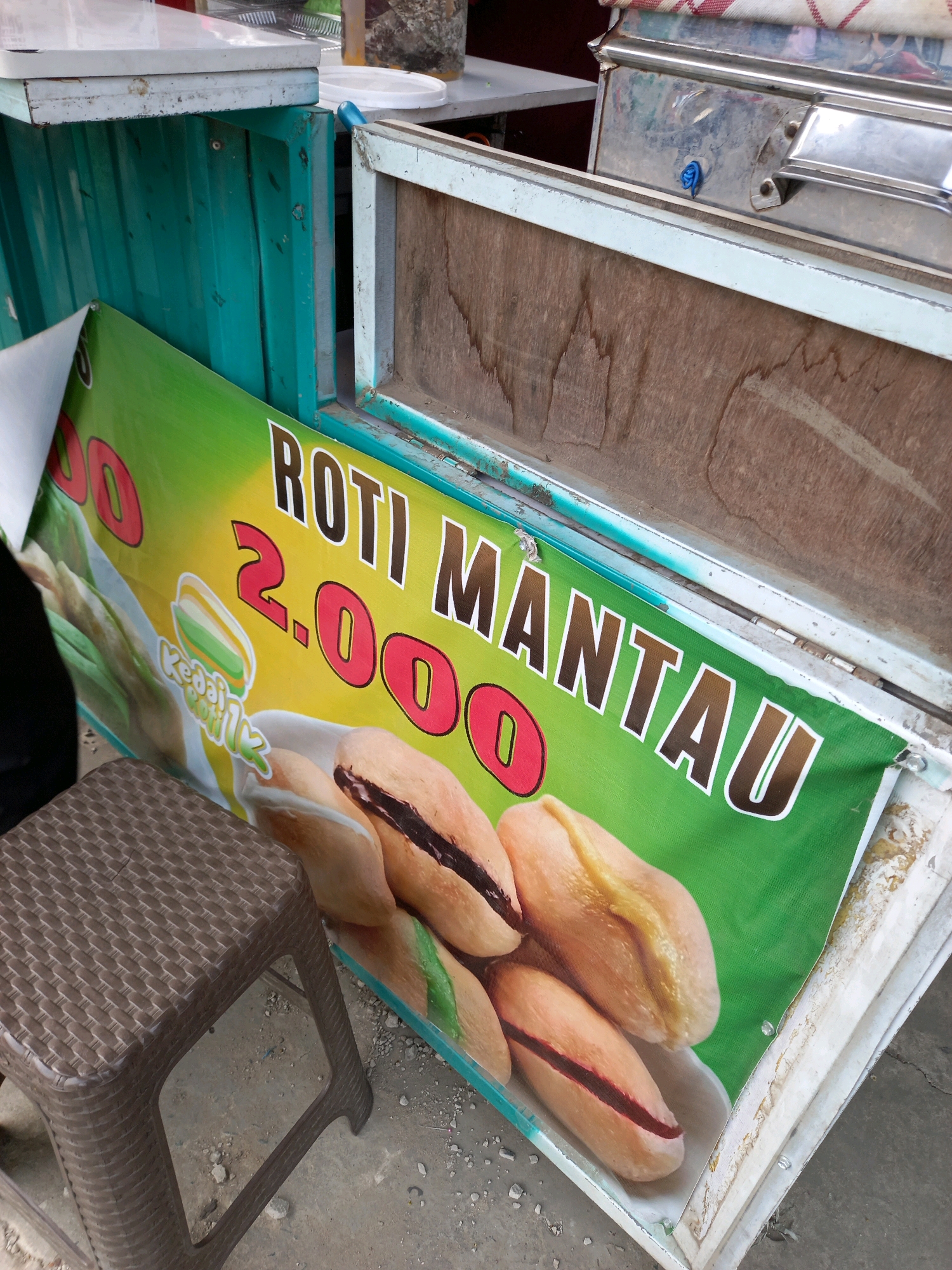 The image shows a close-up of a food stall in Samarinda, Indonesia. A banner with green and yellow gradient displays the text "ROTI MANTAU" and the price "2.000". The banner also features an illustration of bread rolls filled with a dark spread, possibly chocolate or peanut butter. To the left of the banner, a brown plastic stool is visible, placed on a concrete ground with scattered small stones and debris. The stall itself appears to be made of turquoise-colored metal with a wooden interior. Part of a metal cart or food warmer is visible behind the stall. The scene suggests a casual outdoor dining or food vending setting.