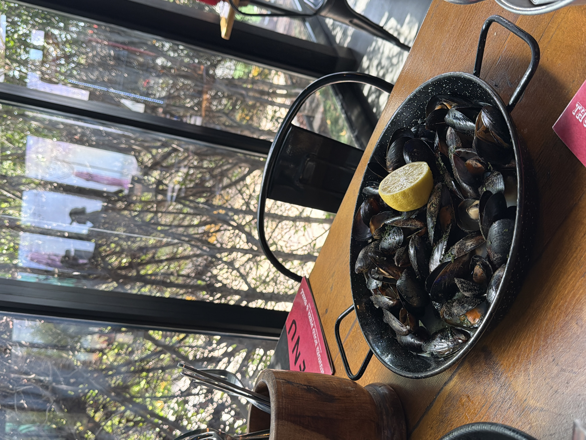 A close-up, eye-level shot shows a serving of cooked mussels in a black pan with handles, garnished with a slice of lemon. The pan sits on a wooden table, with a wooden utensil holder and a red menu to the left. In the background, through large glass windows, bare tree branches and foliage are visible, suggesting an outdoor or garden setting. The scene appears to be an outdoor seating area of a restaurant, likely in Umm Al Quwain, United Arab Emirates, given the natural light and vegetation.
