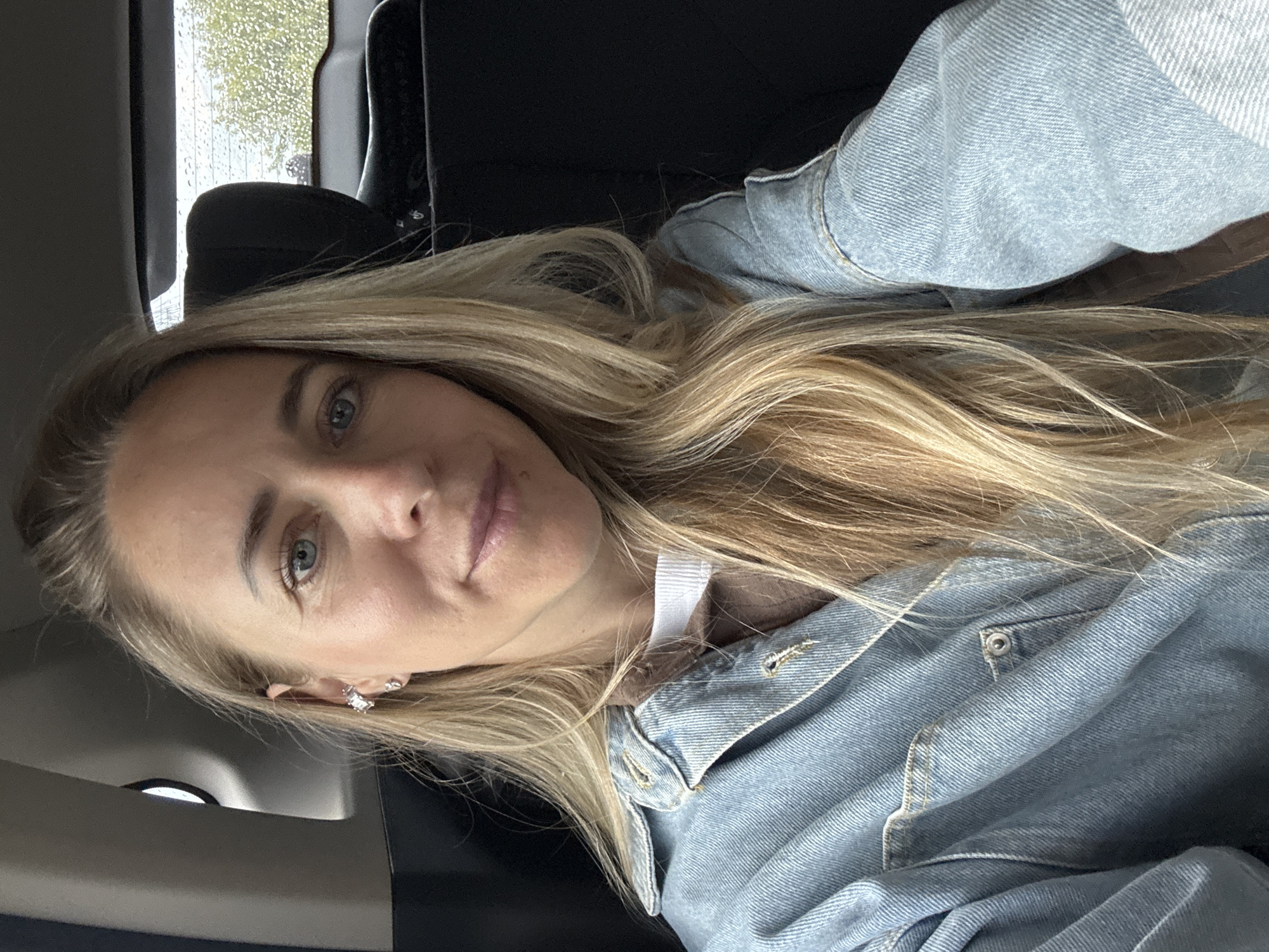 A medium shot selfie shows a woman with blonde hair and blue eyes looking directly at the camera. She is wearing a light blue denim jacket over a white and brown shirt. She has earrings in both ears, one a stud and the other a dangly earring. The woman appears to be sitting in the back seat of a vehicle. The background shows a black car seat and a window with rain droplets on it, revealing greenery outside. There is no information in the image that can definitively place the location in Chaikovsky, Russia.