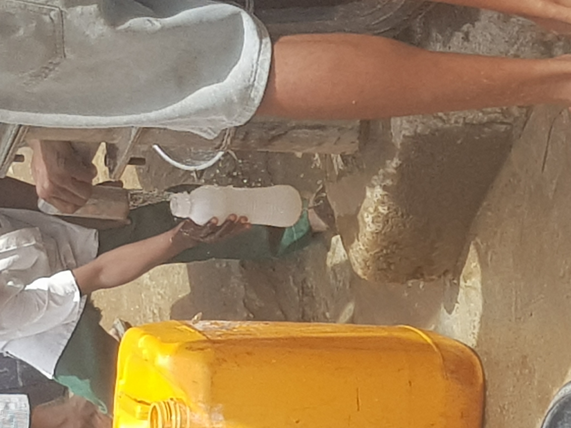 A person wearing a white shirt and green apron fills a white plastic bottle with liquid from a yellow jerrycan in Dutsinma B, Nigeria. The scene includes a rough, light brown surface in the background, with a person's leg and shorts visible in the upper portion of the frame. The jerrycan is in the foreground, taking up a significant portion of the lower image. The focus is on the action of pouring the liquid.