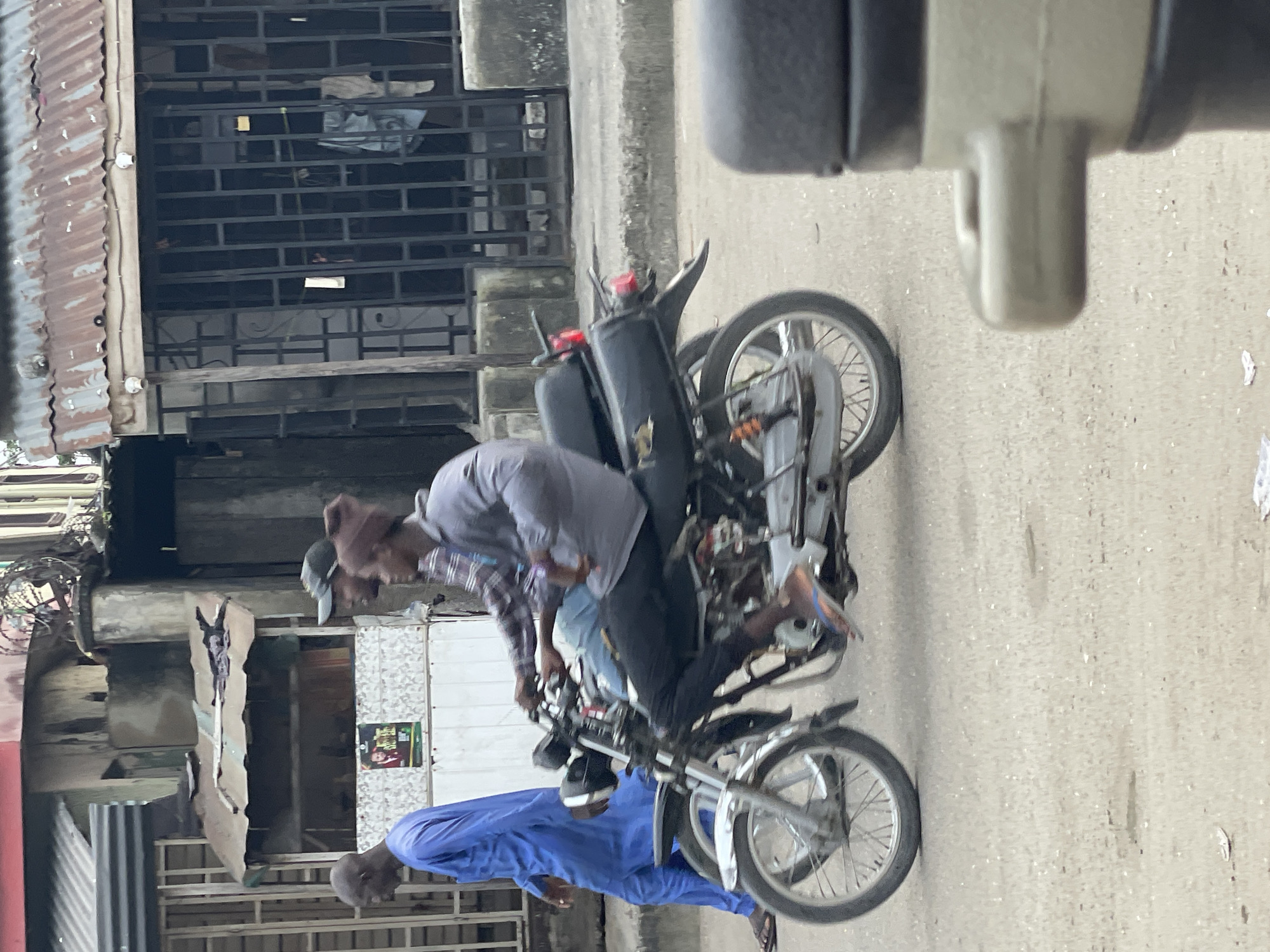 The image depicts a street scene in Ado, Nigeria, with a motorcycle carrying two men in the foreground. The motorcycle is black and silver with a visible rear tail light and tire. The man driving is wearing a grey long-sleeved shirt and dark pants, with a brown beanie on his head. The passenger behind him is wearing a checkered shirt and a dark baseball cap. To the left of the motorcycle, a man in a bright blue robe is walking away from the camera. In the background, there are buildings with corrugated metal roofs and barred windows, suggesting a commercial or residential area. The ground is paved, appearing to be a road or sidewalk.
