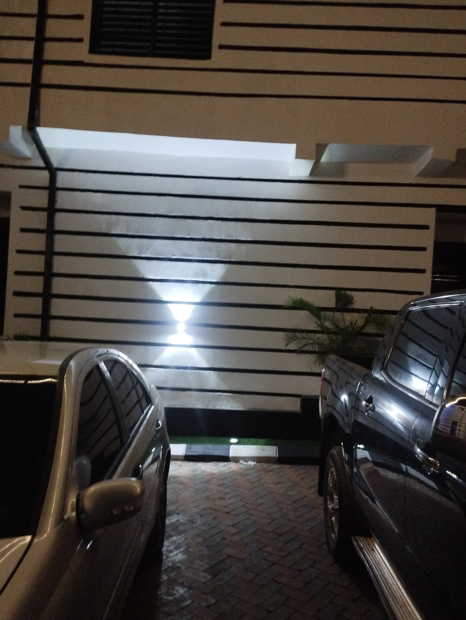 The image shows a nighttime scene in Tumfure, Nigeria. A light-colored car is parked on the left, and a dark pickup truck is parked on the right. Behind the vehicles is a building with horizontal black lines on a light-colored wall. A light fixture is mounted on the wall between the vehicles. A small plant is visible between the truck and the wall. The ground appears to be paved with bricks.