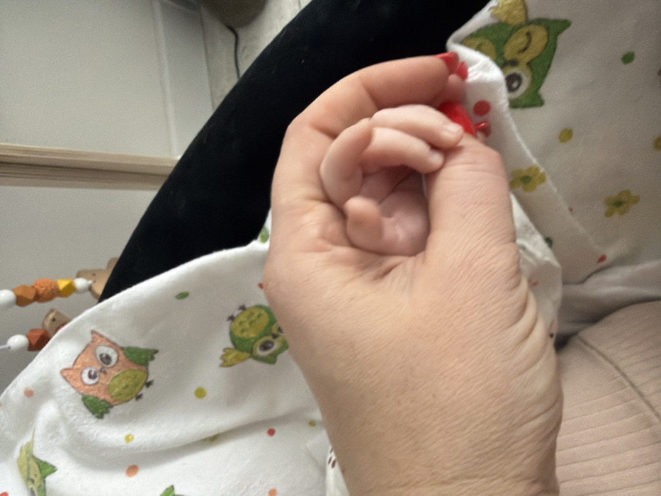 A close-up shows an adult hand with red nail polish gently holding a baby's tiny hand. The baby's hand is curled in a small fist. The adult's arm is visible, showing skin texture. The background is a white blanket with a pattern of cartoon owls and colorful dots and flowers. A black object and a wooden toy with beads are visible in the blurred background.