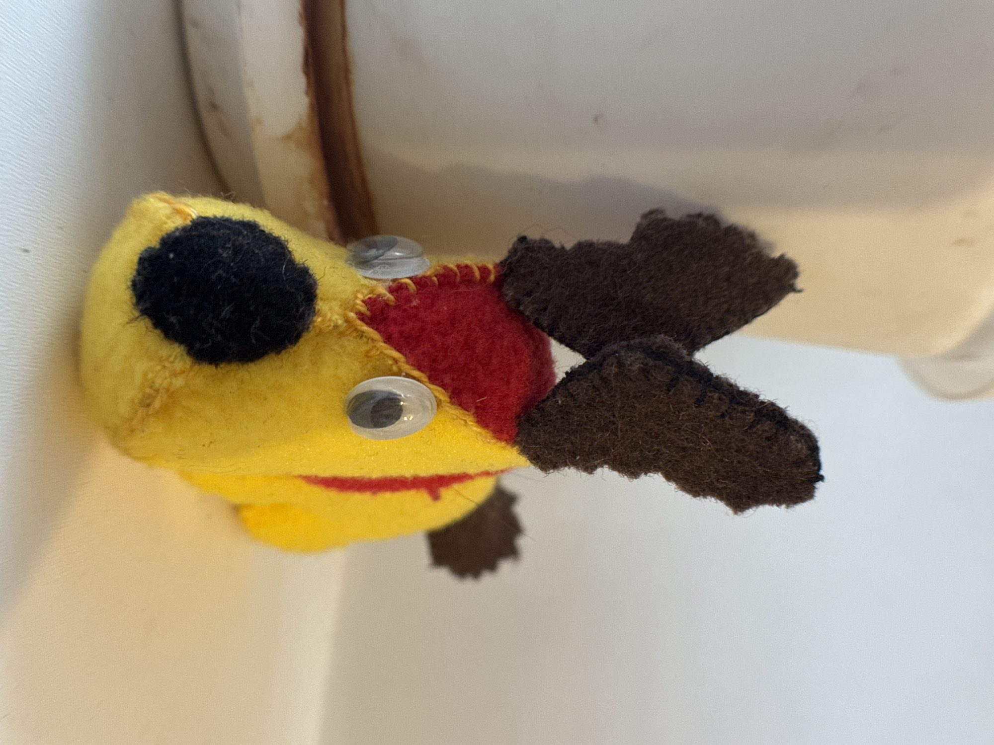 A handmade, colorful toy is positioned against a light-colored backdrop. The toy has a yellow head with a black circle stitched onto its left side. It has two plastic eyes and a red segment stitched to the yellow head. The toy has dark brown flipper-like appendages. The context of the city of Novo-Nikol'skoe, Russia, is not applicable based on the visual information in the image.