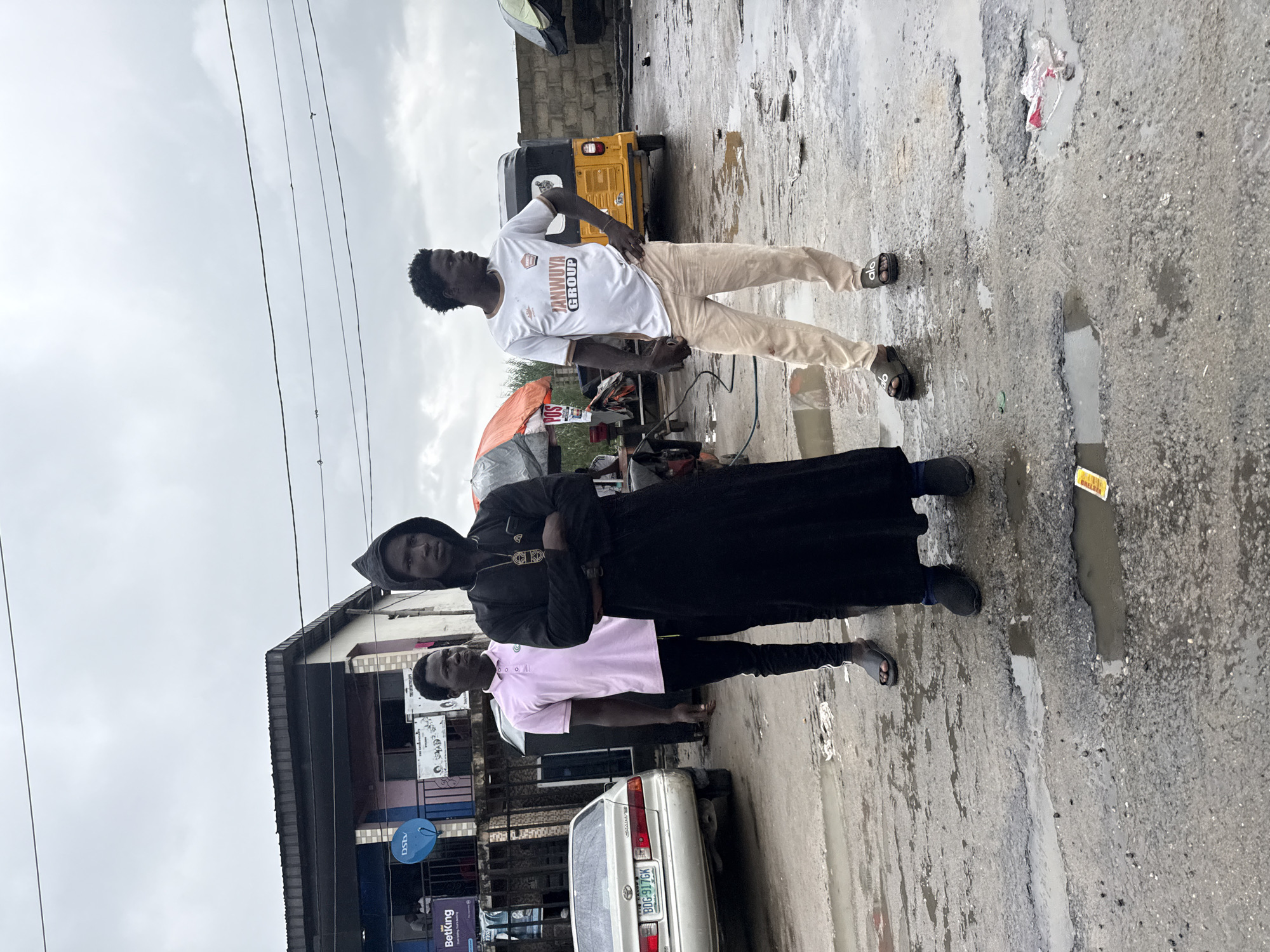 In Ado, Nigeria, under a cloudy sky, three individuals stand on a wet, pothole-ridden asphalt surface. To the left, a silver sedan is parked. In the center, a person in a pink polo shirt and black pants stands next to someone in a black hooded robe. To the right, a man in a white t-shirt and light-colored pants is positioned near a yellow auto-rickshaw. Various stalls and buildings, including one with a DStv satellite dish, are visible in the background.