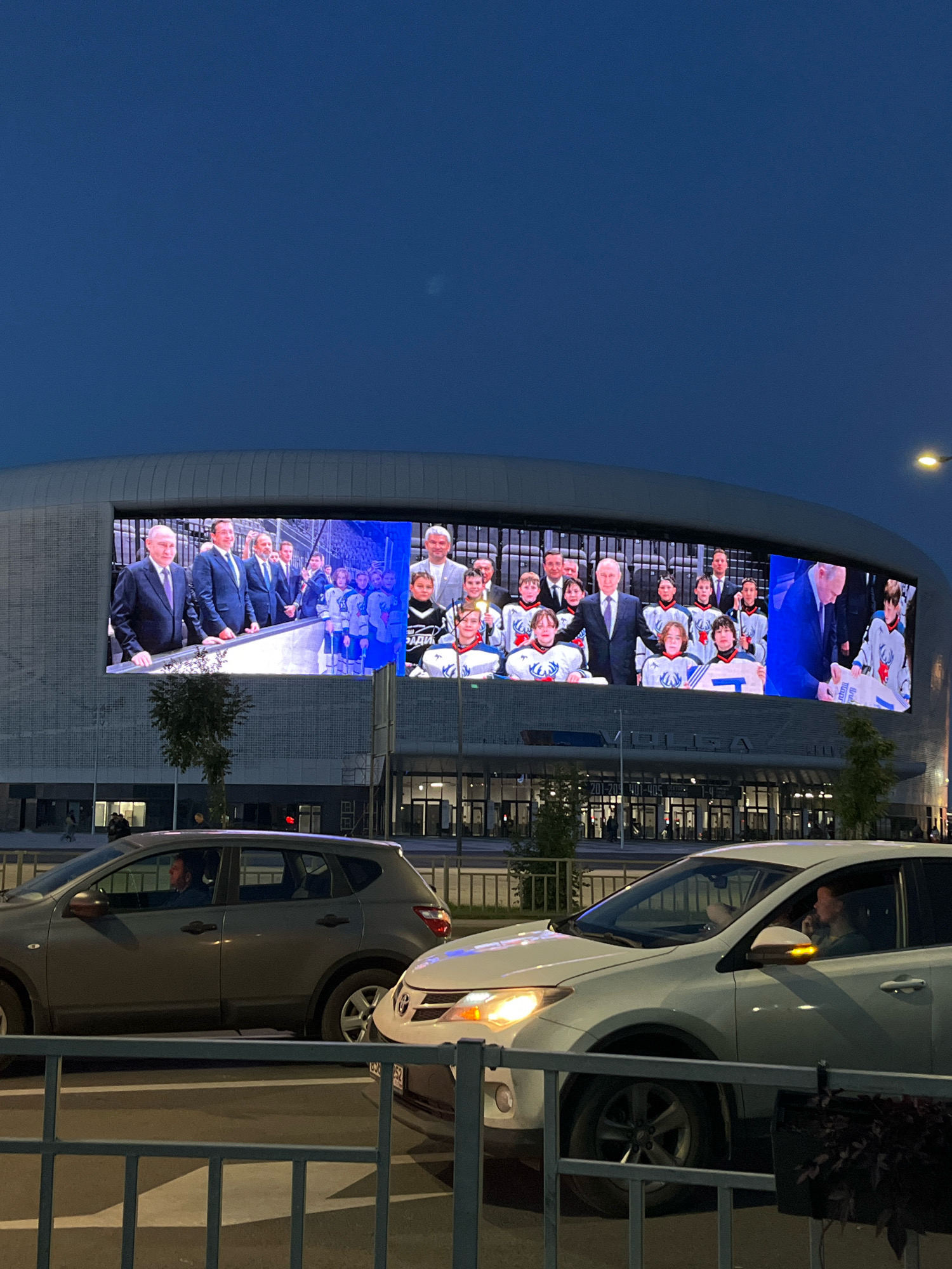 The image captures a view of the Nizhny Novgorod Stadium at dusk. A large LED screen on the stadium facade displays a scene of dignitaries in suits interacting with a group of young people in hockey uniforms. The sky above is a deep blue, characteristic of twilight. In the foreground, two cars are parked or stopped, with the occupants visible inside. The overall setting appears to be an urban environment, with the stadium serving as a prominent landmark.