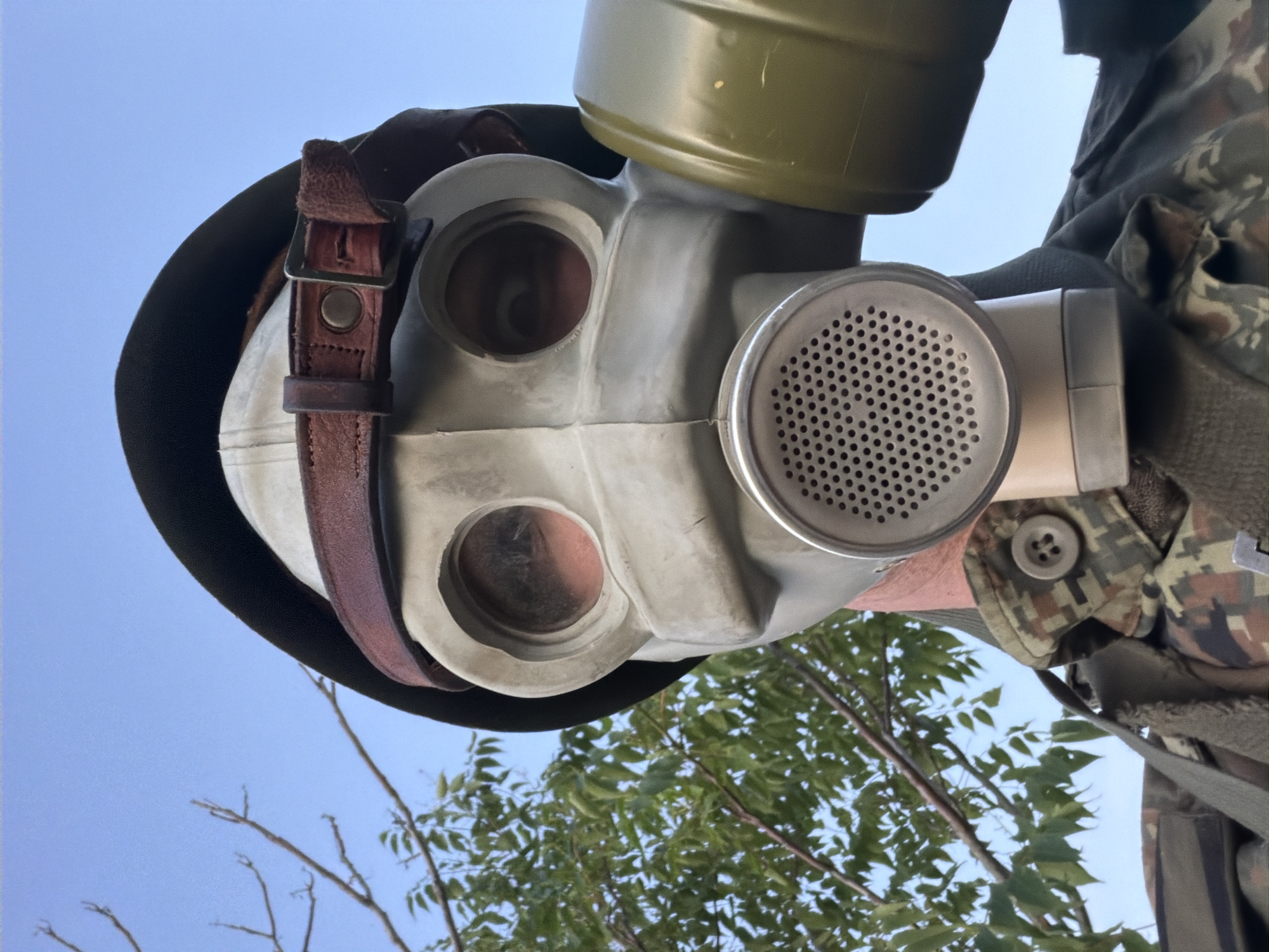A person wearing a gas mask and military uniform is shown from the chest up, outdoors with a clear blue sky and trees in the background. The gas mask is off-white with two large eye lenses, a filter on the right side, and a prominent circular exhalation valve. A brown leather strap is fastened across the top of the mask and under a dark cap. The uniform is camouflage patterned with a button visible on the collar. The person is looking directly at the camera. The location is identified as Samovodene, Bulgaria.