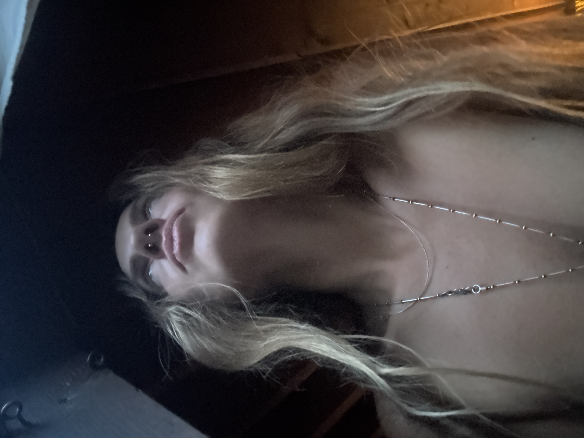 A low-angle, medium shot captures a person with blonde hair looking upwards in an indoor environment. The person has a nose ring and is wearing two necklaces. One necklace is a thin silver chain, and the other is a longer silver chain with small beads and a pendant. The background is dark and appears to be a ceiling or overhead structure. The lighting is dim, with warm tones illuminating the right side of the person's face and hair.