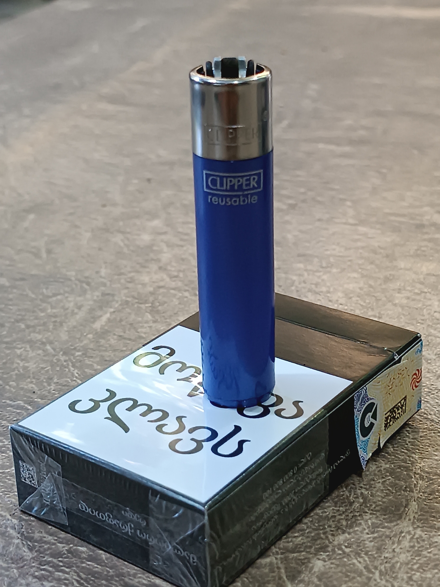 A blue Clipper lighter with "CLIPPER reusable" printed in white stands upright on a pack of cigarettes. The cigarette pack has a black border and a white top with large, stylized Georgian script in a dark green color. The side of the pack displays colorful patterns and some text in Georgian. The scene is set on a textured, gray surface, likely a table. There are no people visible in the image. This scene appears to be a still life of smoking paraphernalia.