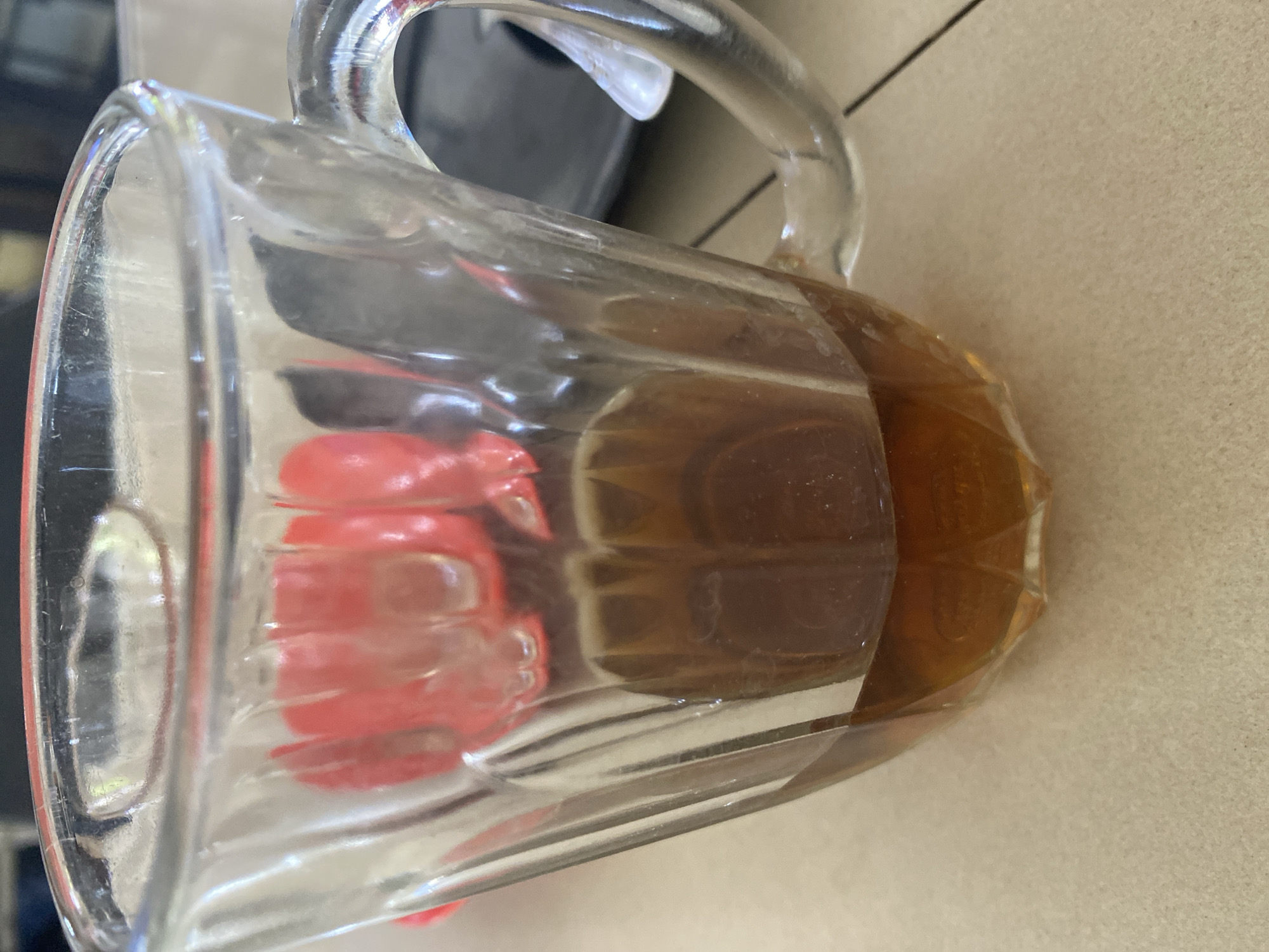 A close-up, eye-level shot shows a glass mug with a handle containing a dark amber liquid, likely tea. The mug has vertical ridges on its exterior and is situated on a speckled beige surface, possibly a table. The liquid fills approximately one-third of the mug. In the background, out of focus, a dark plate or tray is visible, and behind that, a pink object with an irregular shape can be discerned. The lighting suggests an indoor setting, possibly a cafe or home, in Lhokseumawe, Indonesia. No individuals are present in the frame.